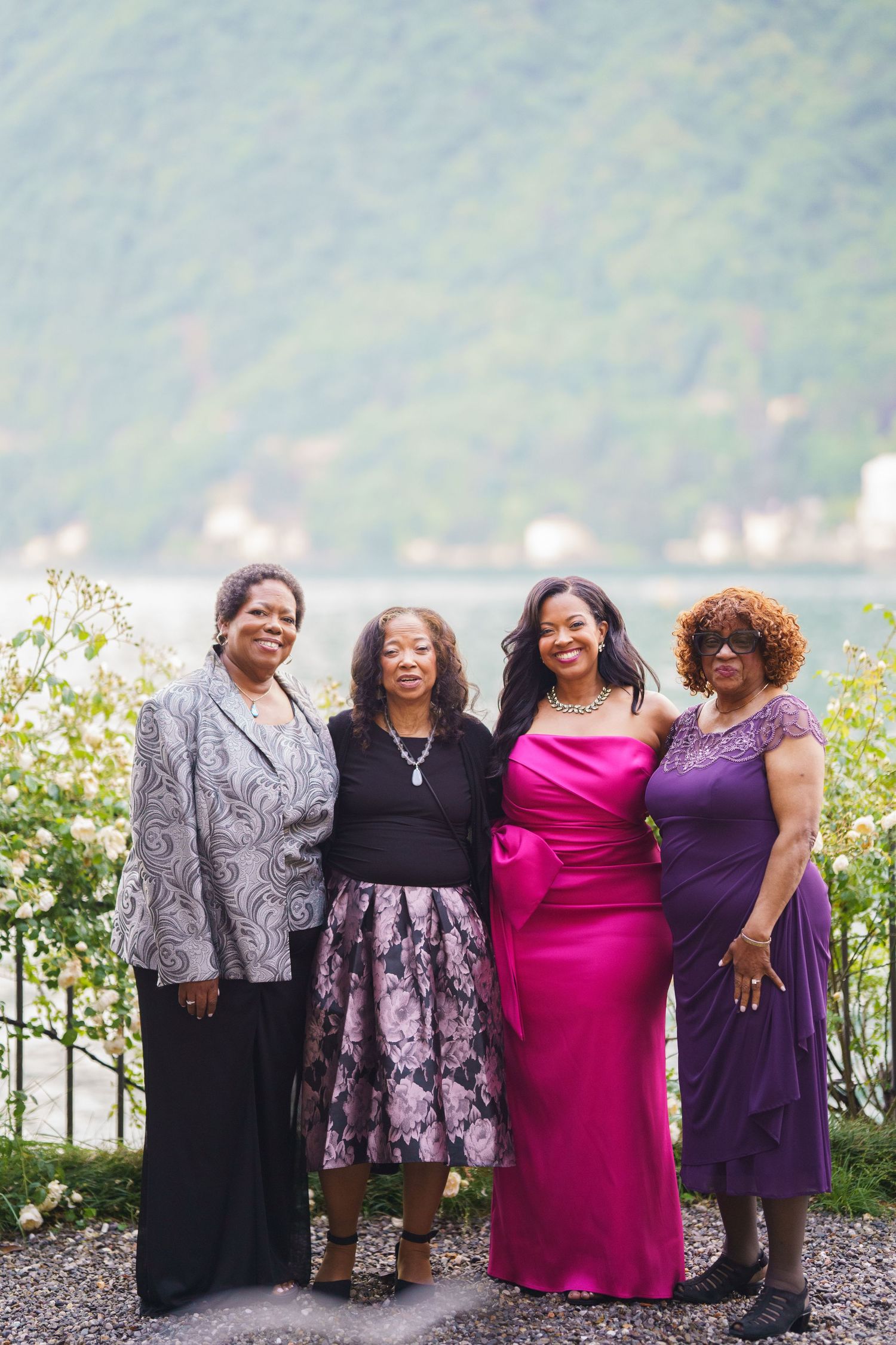 Four women in elegant dresses pose together outdoors near a lake with lush green hills in the background.