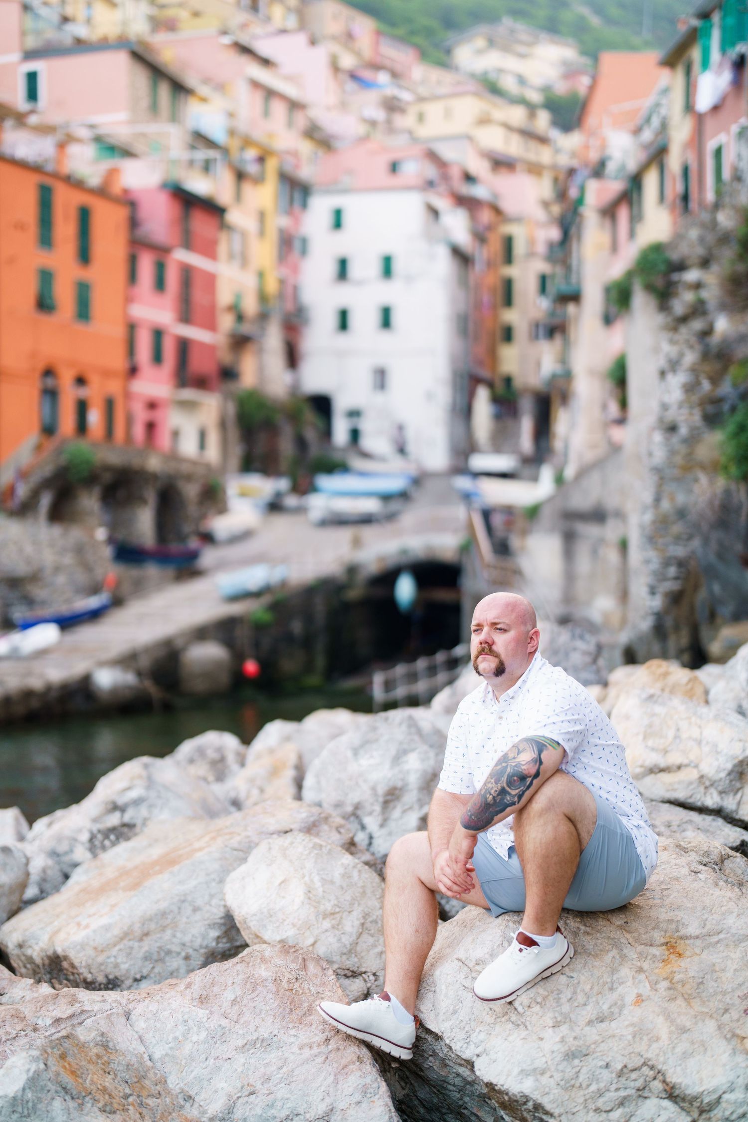 Bald tattooed man sitting on rocks in Riomaggiore, Cinque Terre, Italy, with colorful buildings and boats in background.