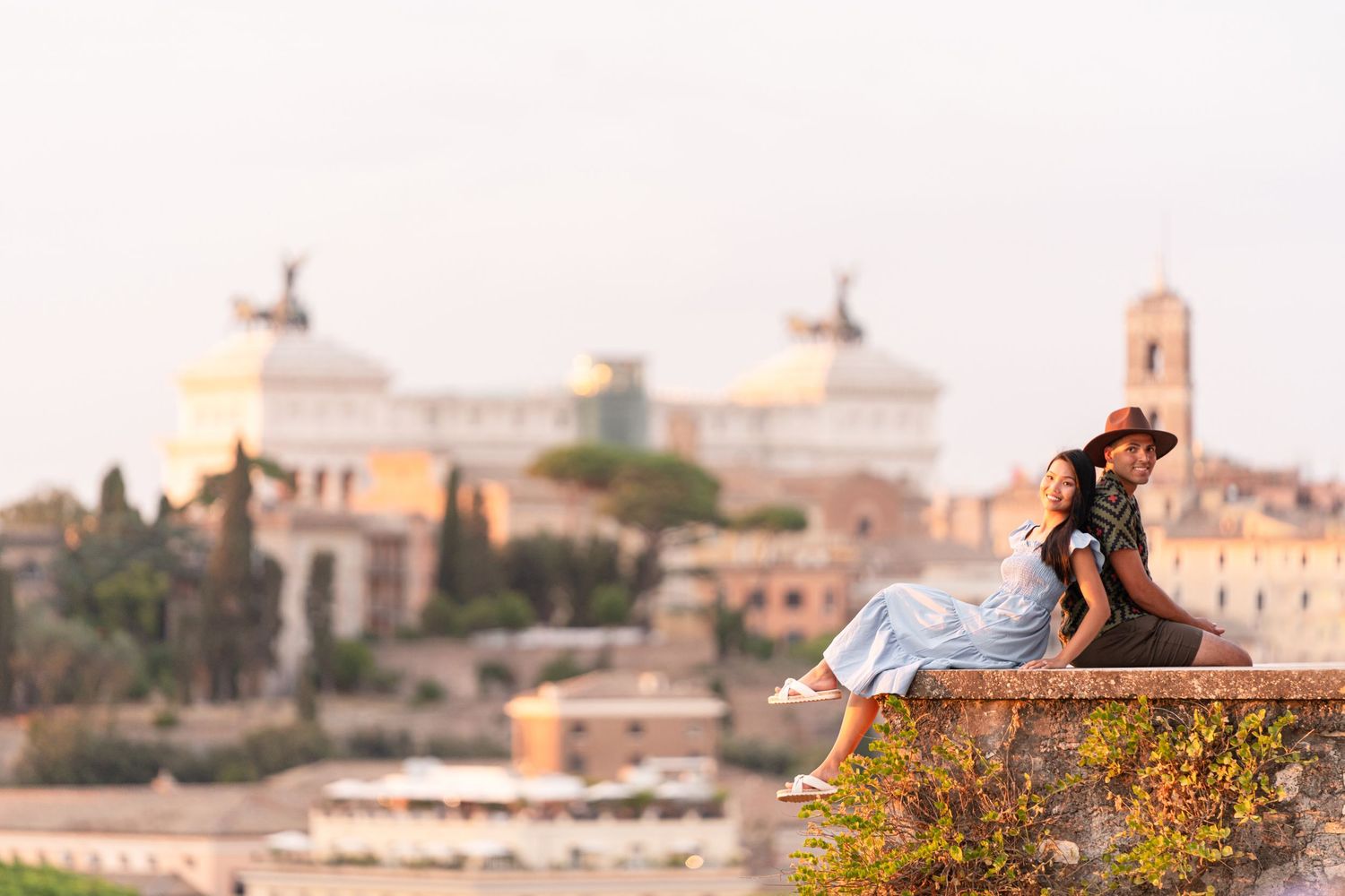 Couple sitting on ancient stone wall with Rome skyline and Vittoriano monument in background at sunset.
