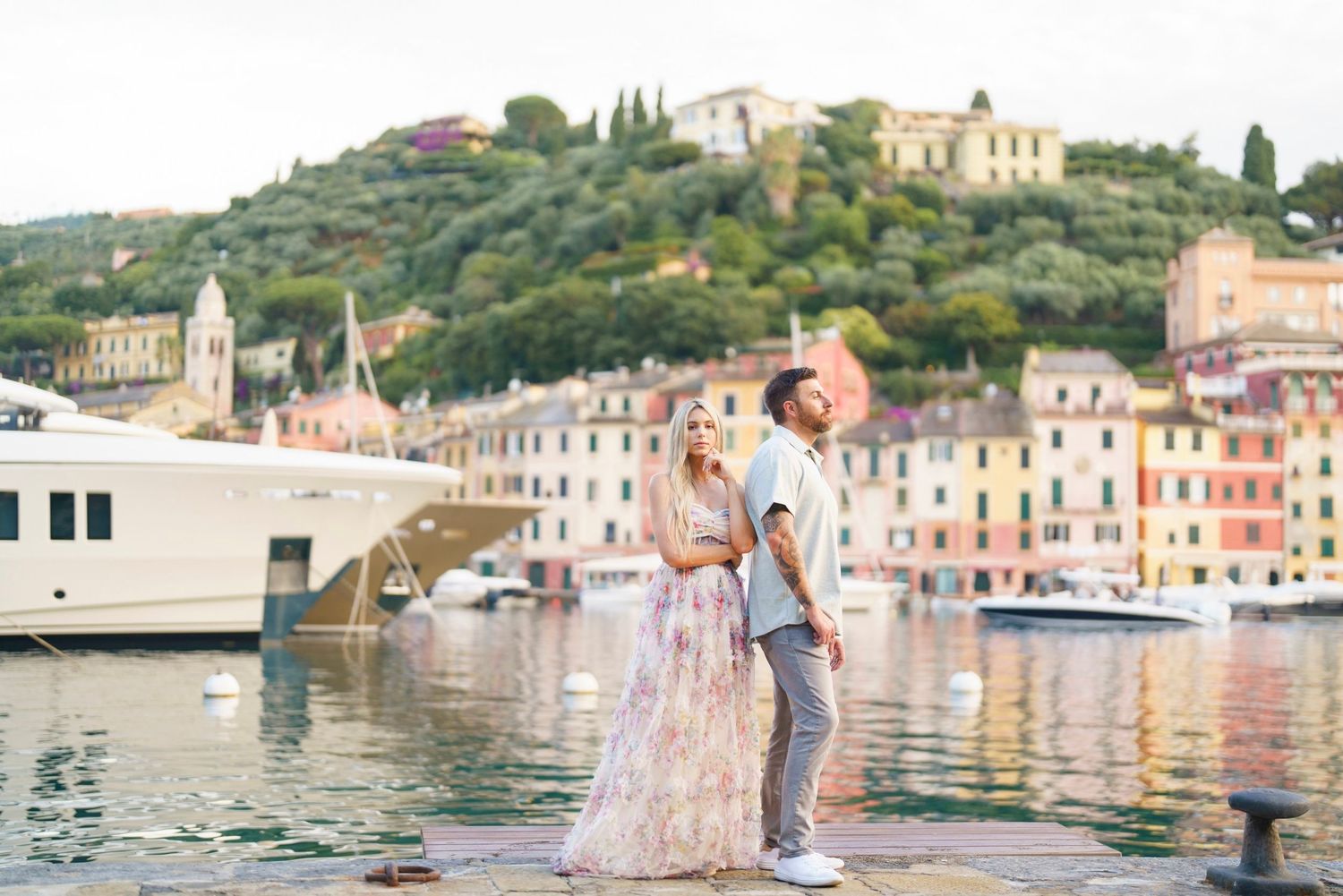 Couple posing on a dock in Portofino, Italy, with colorful buildings and a luxury yacht in the background.