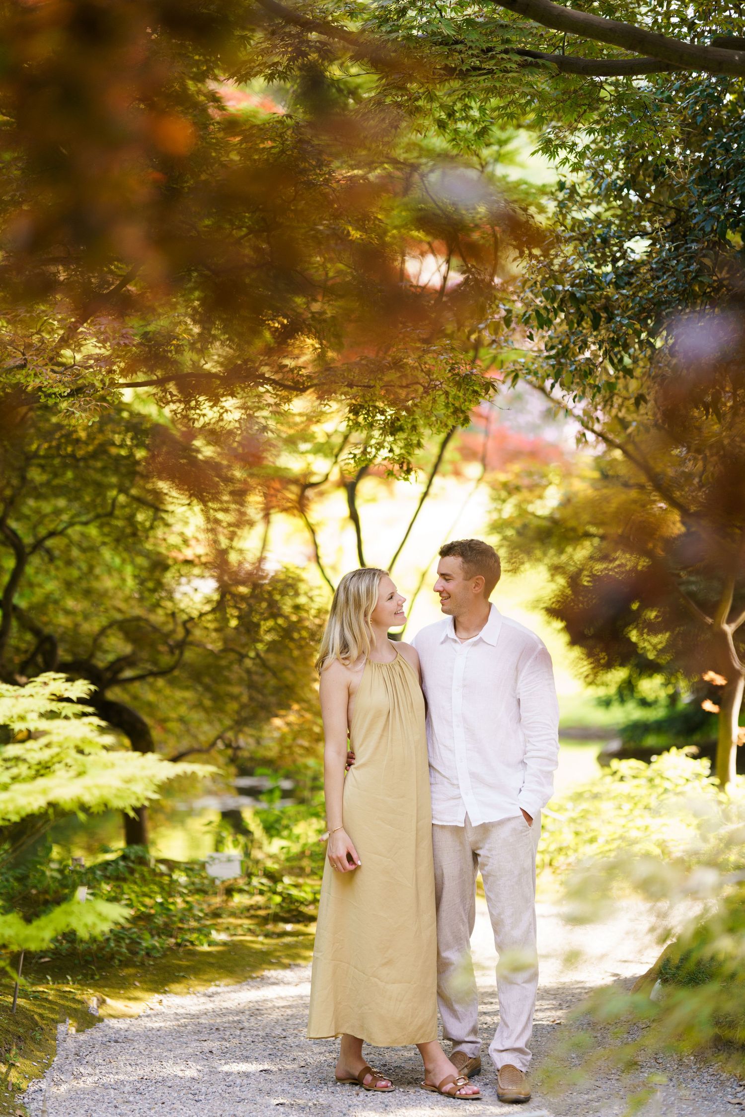 Couple in a lush garden path, woman in yellow dress, man in white shirt, surrounded by colorful autumn trees.