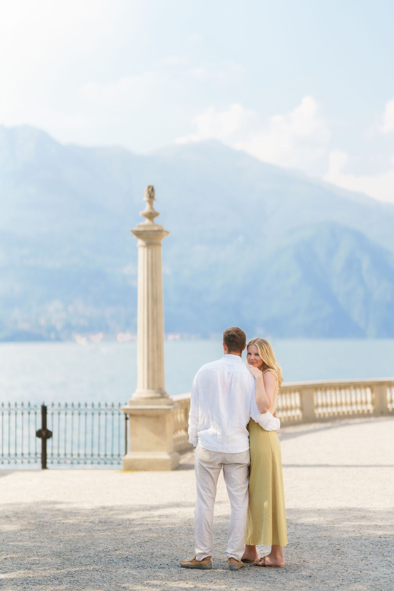 Couple embracing on a scenic lakeside terrace with mountains and blue sky in the background.