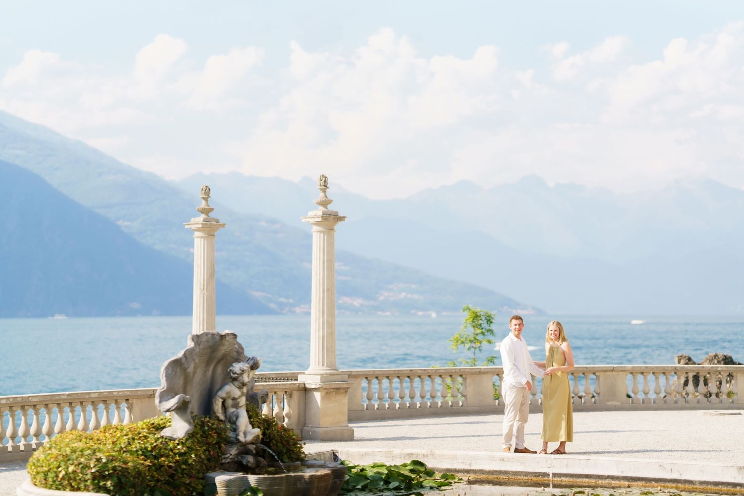 Couple holding hands on elegant lakeside terrace with marble columns, fountain, and stunning mountain backdrop.