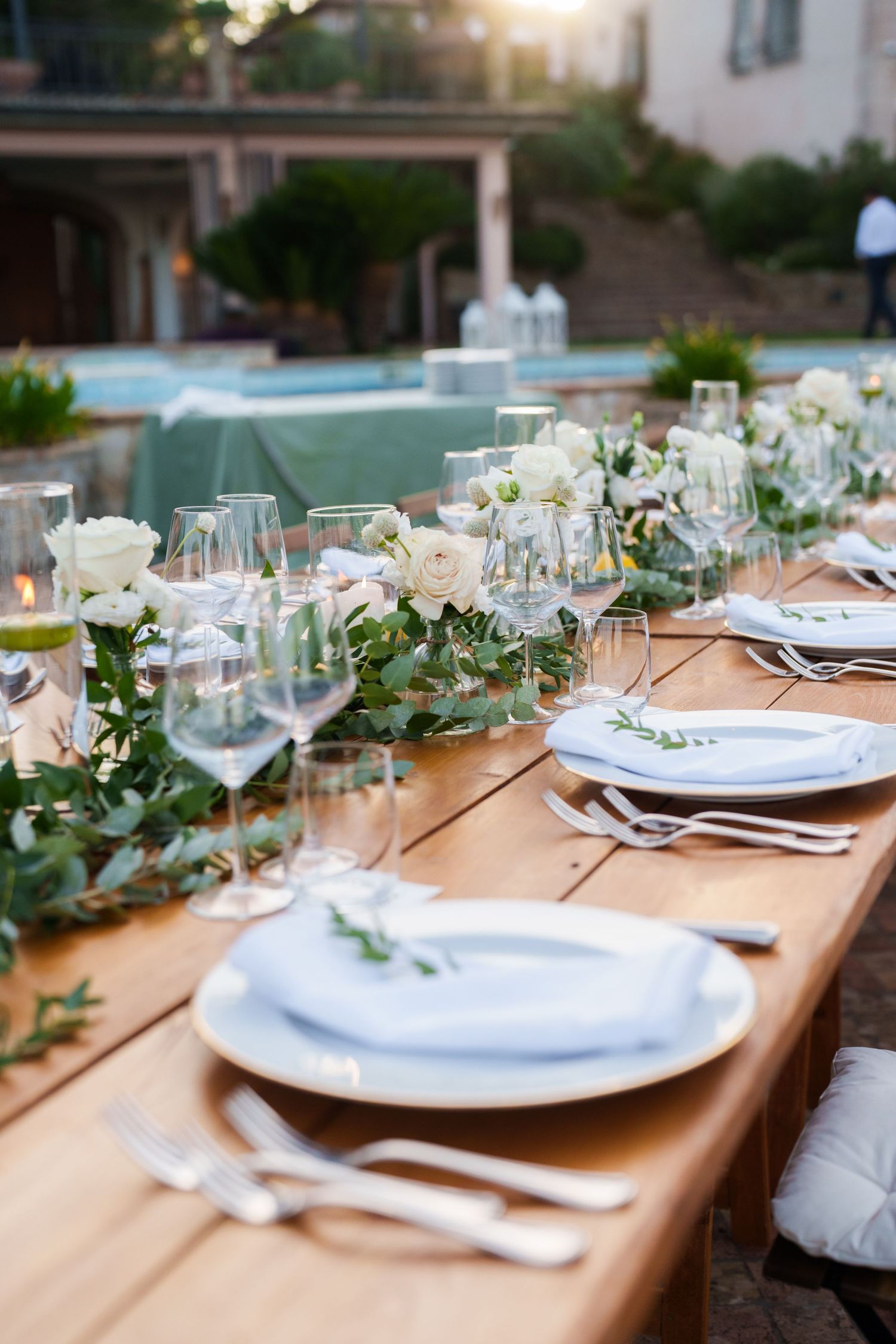 Elegant outdoor wedding reception table with white roses, greenery garland, wine glasses, and white plates by a pool.