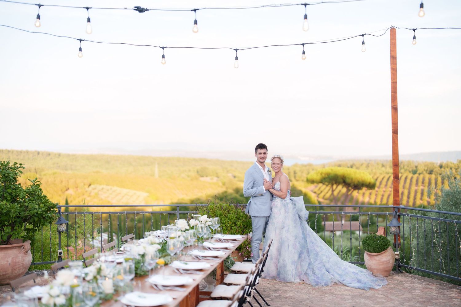 Bride and groom pose at elegant outdoor vineyard wedding reception with long table, string lights, and scenic Tuscany views.