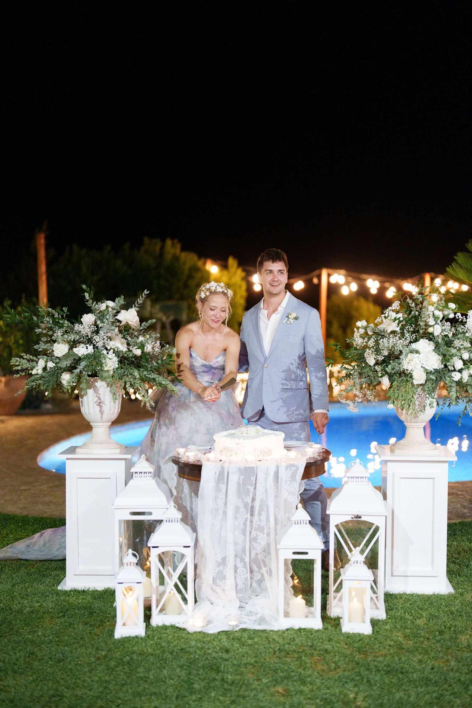 Bride and groom stand at elegant white cake table with lanterns and floral arrangements by a pool at night.