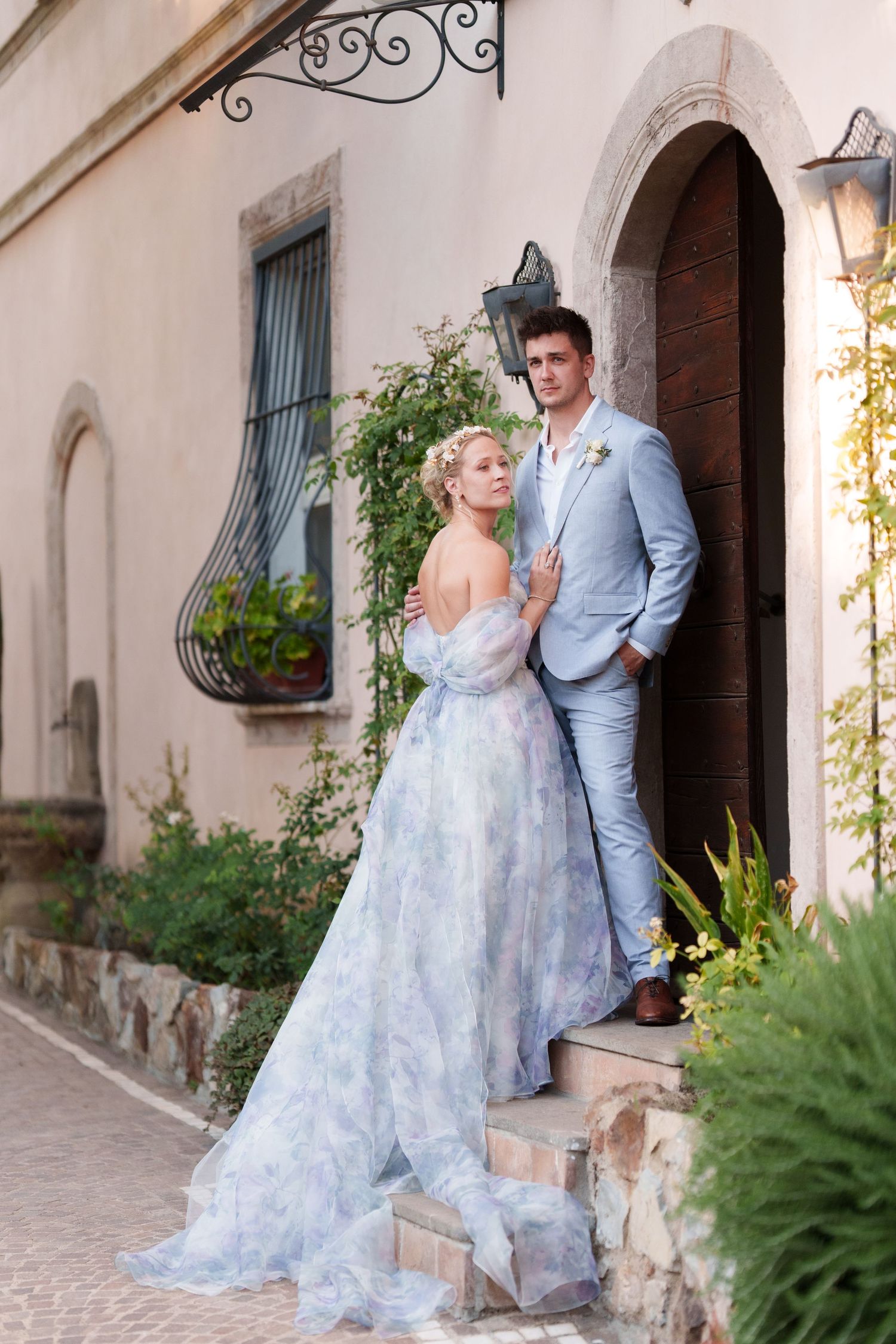 Couple in elegant wedding attire pose by rustic stone building, bride in blue floral gown, groom in light blue suit.