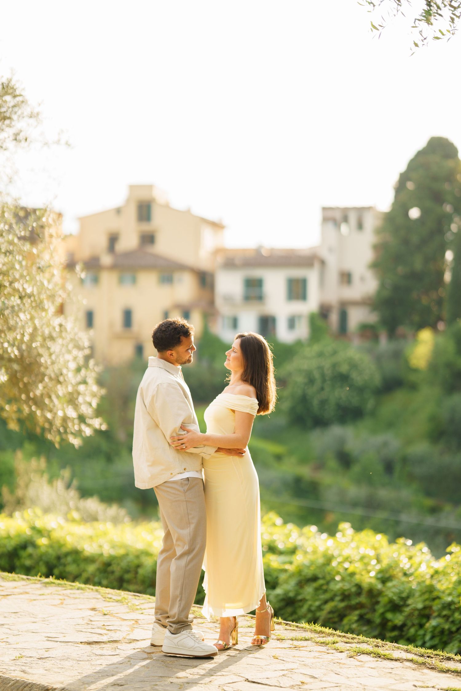 Couple embracing in golden sunlight with Italian architecture and lush greenery in the background.