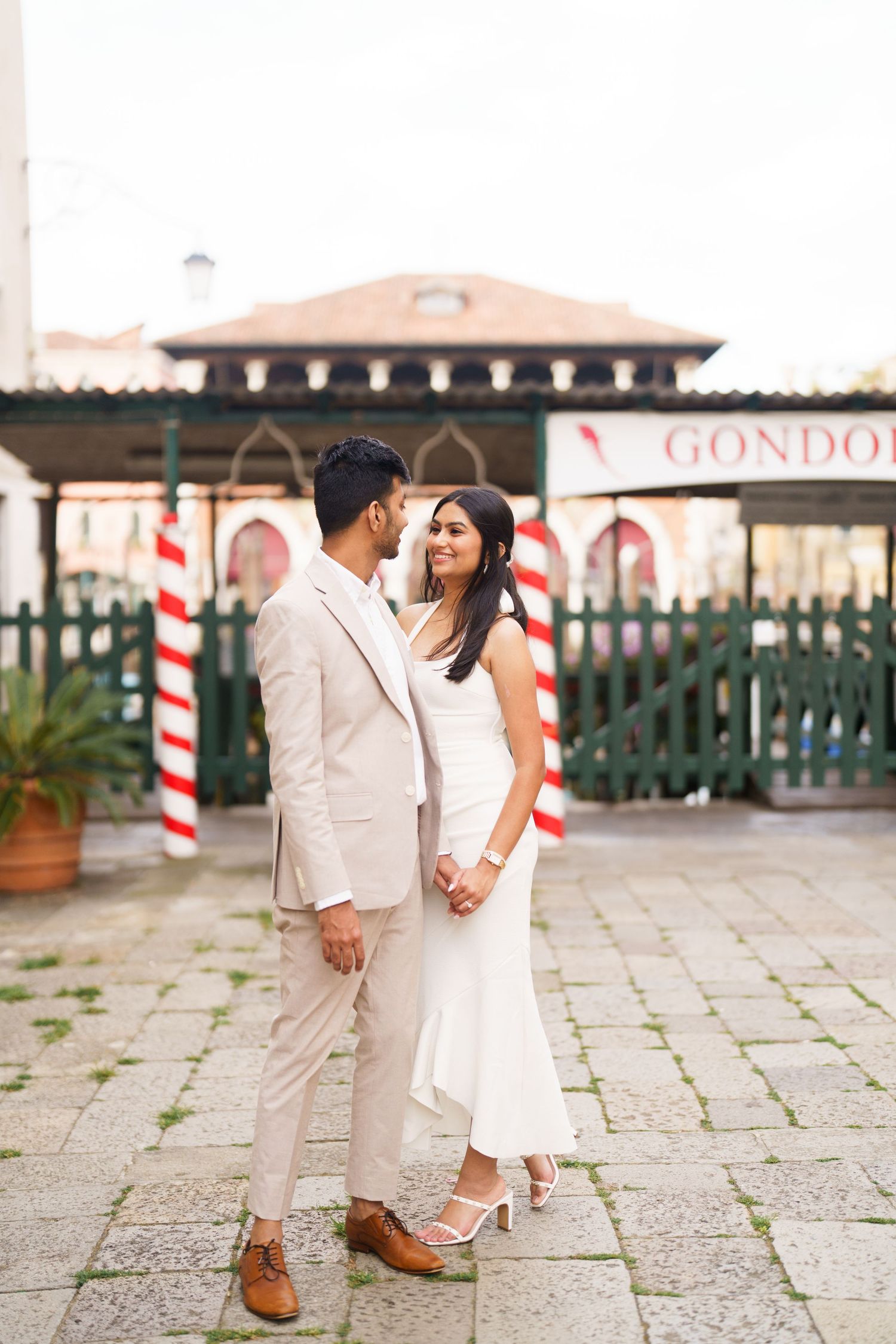 A romantic couple in white outfits stands on a stone patio near a gondola station in Venice, Italy.