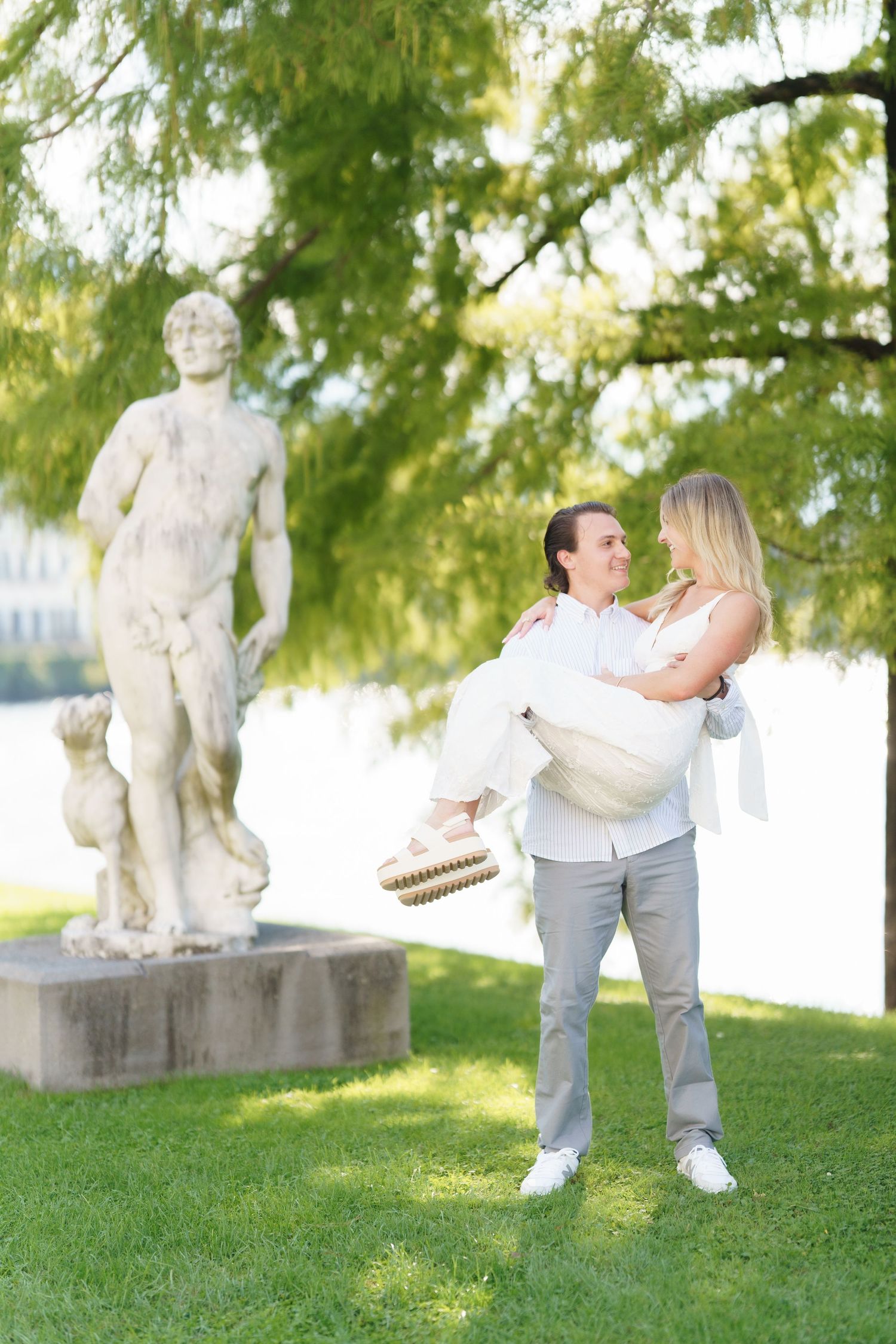 A couple in white outfits playfully poses together on a grassy lawn with stone statues in the background.