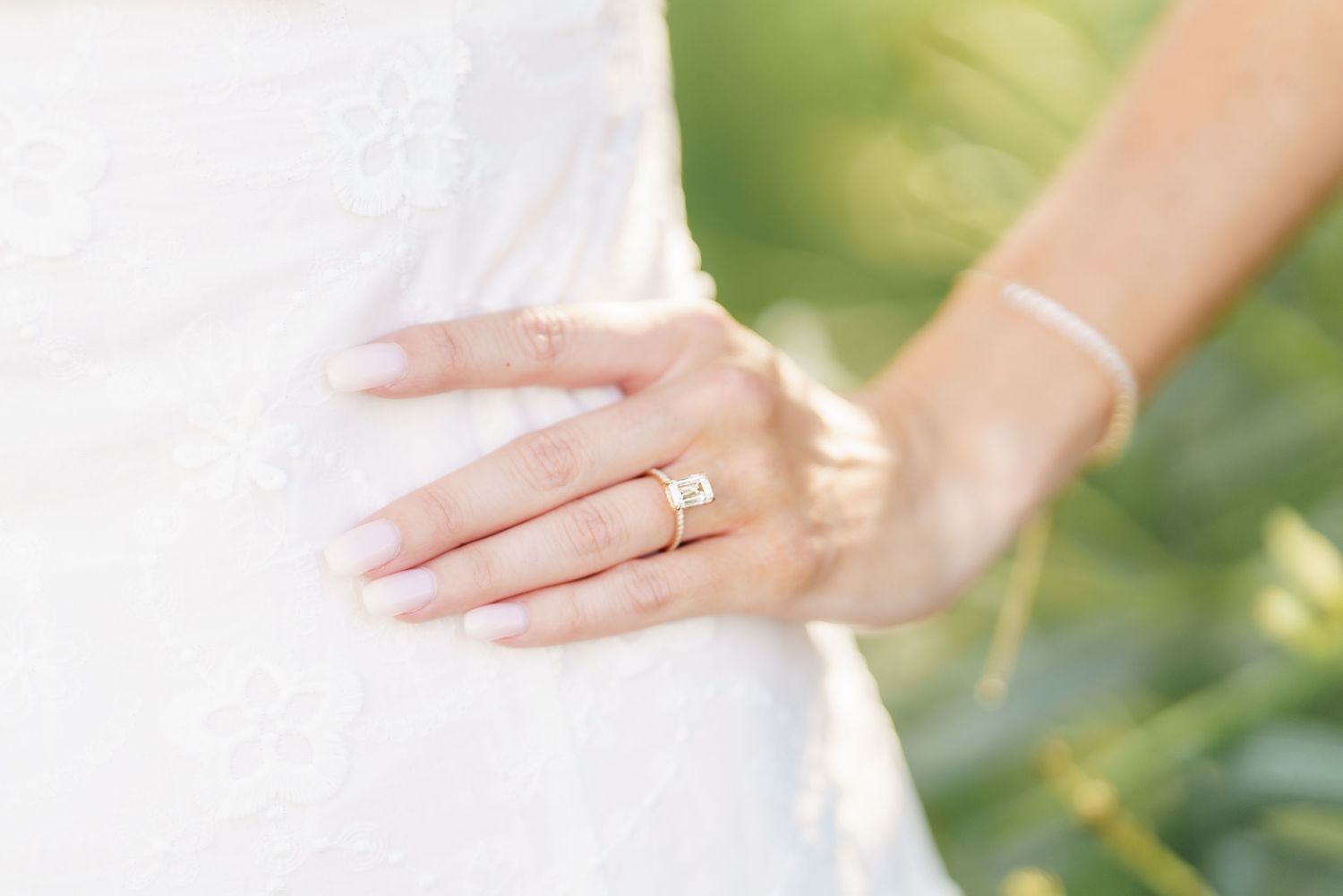 A delicate hand wearing a gold wedding ring rests gently against white fabric in soft natural lighting.