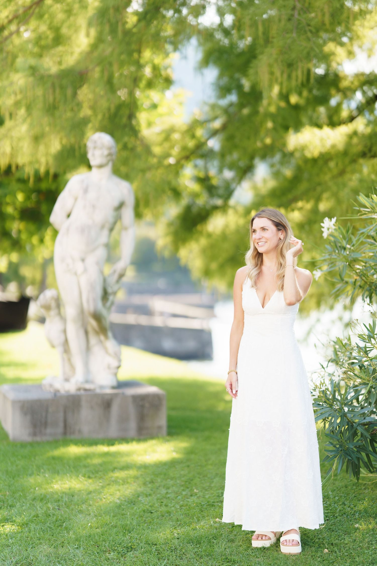 A person in a flowing white dress walks through a sunlit garden with stone sculptures and lush greenery.