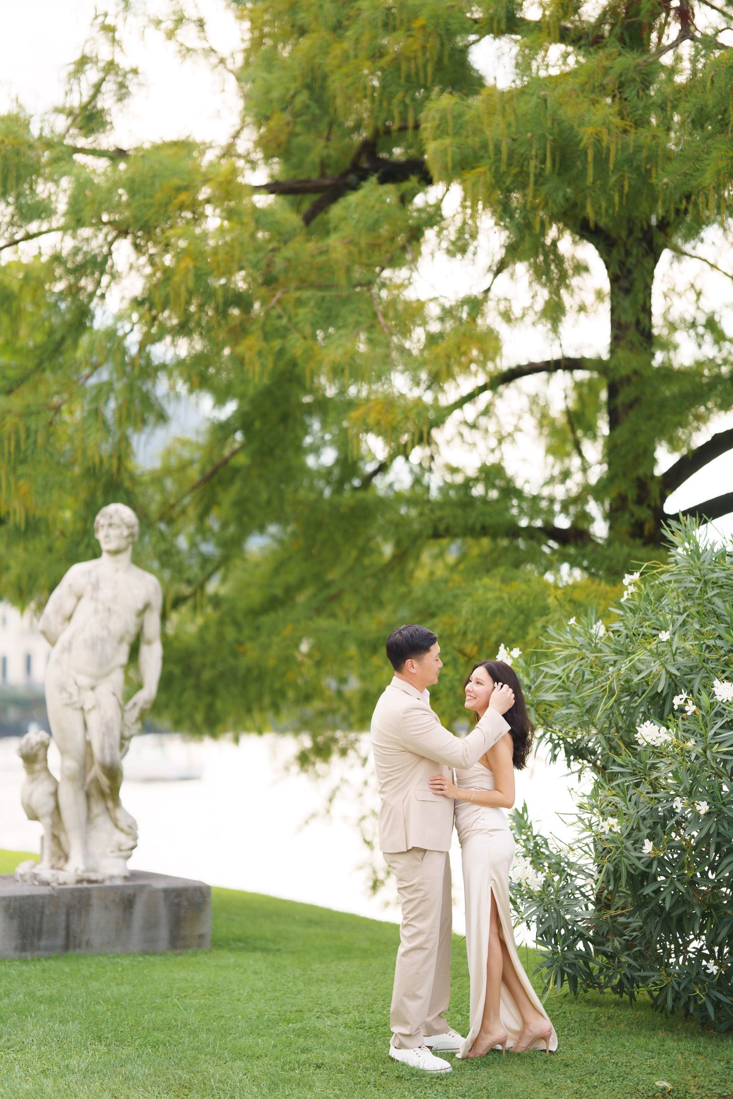 A romantic engagement photoshoot in a garden with classical statues and lush greenery in the background.