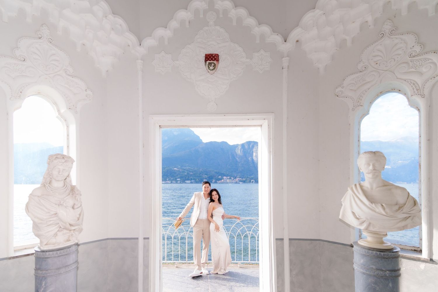 A couple embraces in a white arched terrace overlooking the Amalfi Coast in Italy with classical marble busts on display.