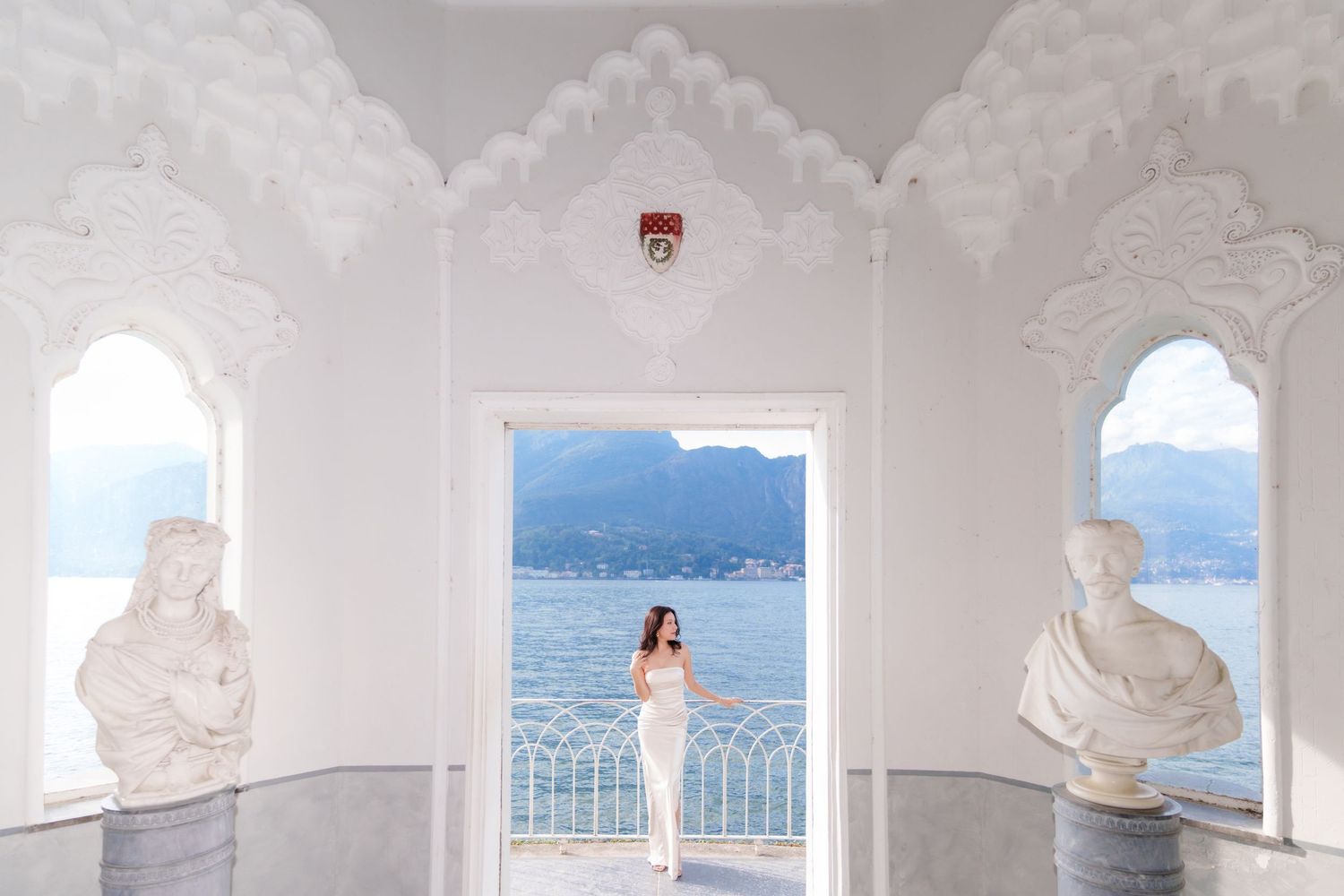 White arched colonnade with ornate architectural details overlooking a scenic mountain lake view in Italy.