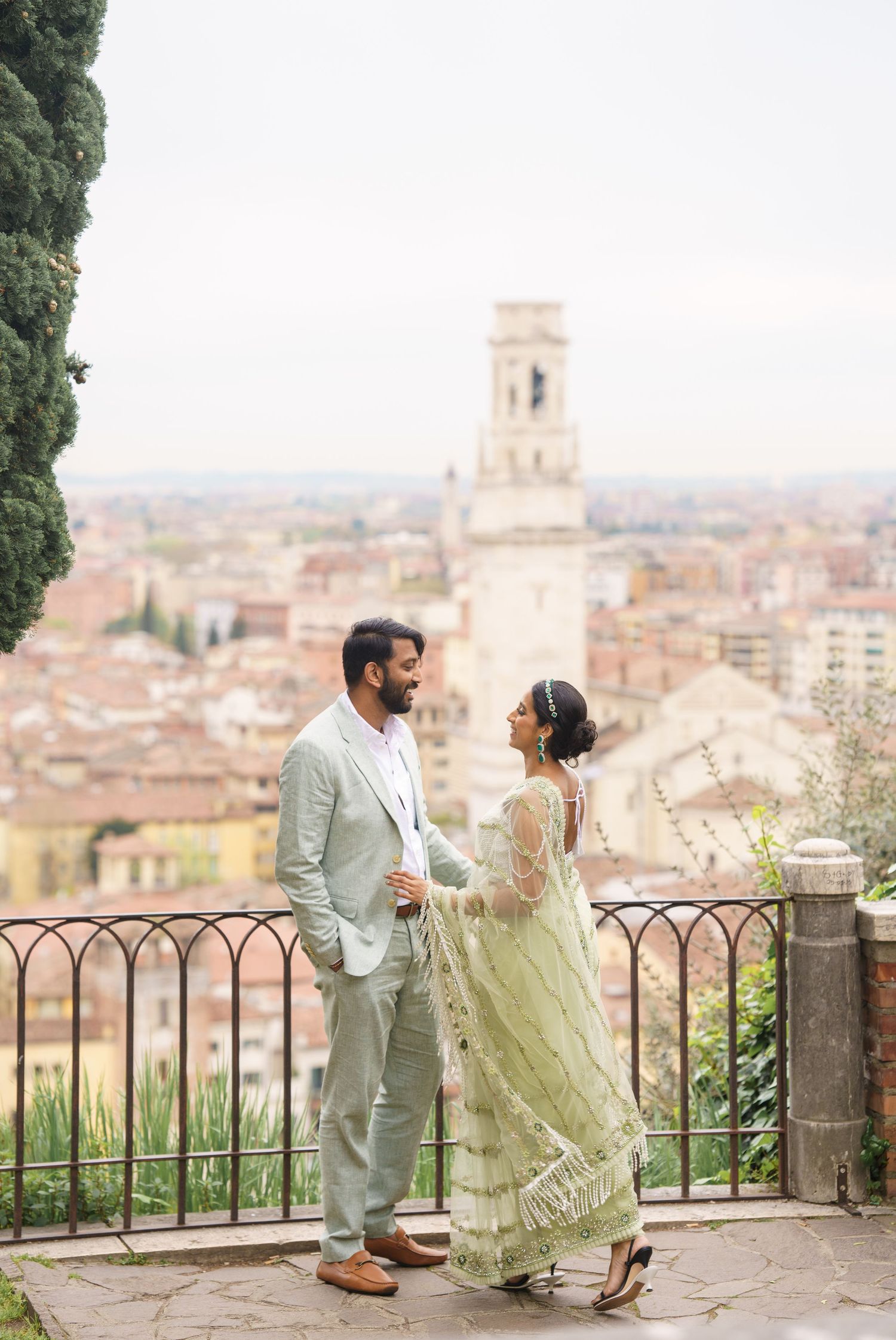 A couple shares a romantic moment at a scenic overlook with a historic Italian cityscape and bell tower in the background.
