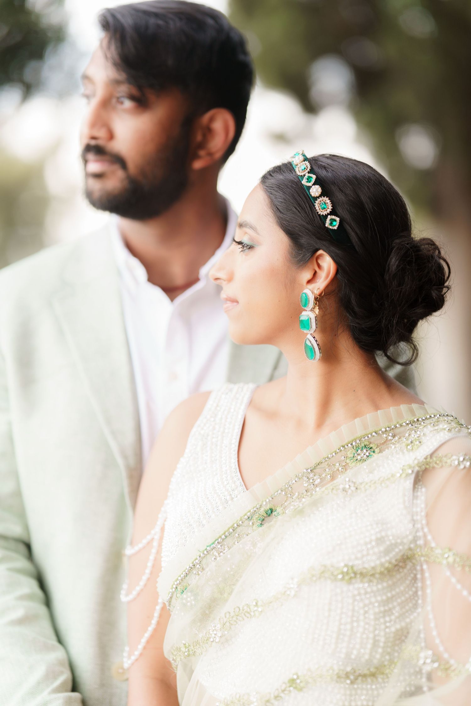 A couple in traditional Indian wedding attire pose together, with the bride wearing emerald earrings and cream saree.