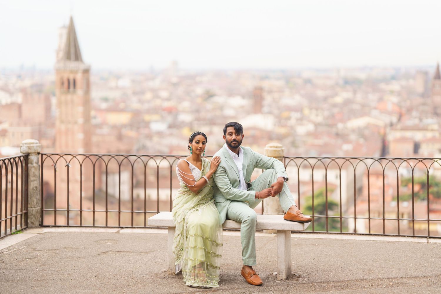 Couple in formal attire enjoying a romantic moment at a scenic overlook with a European city skyline in the background.