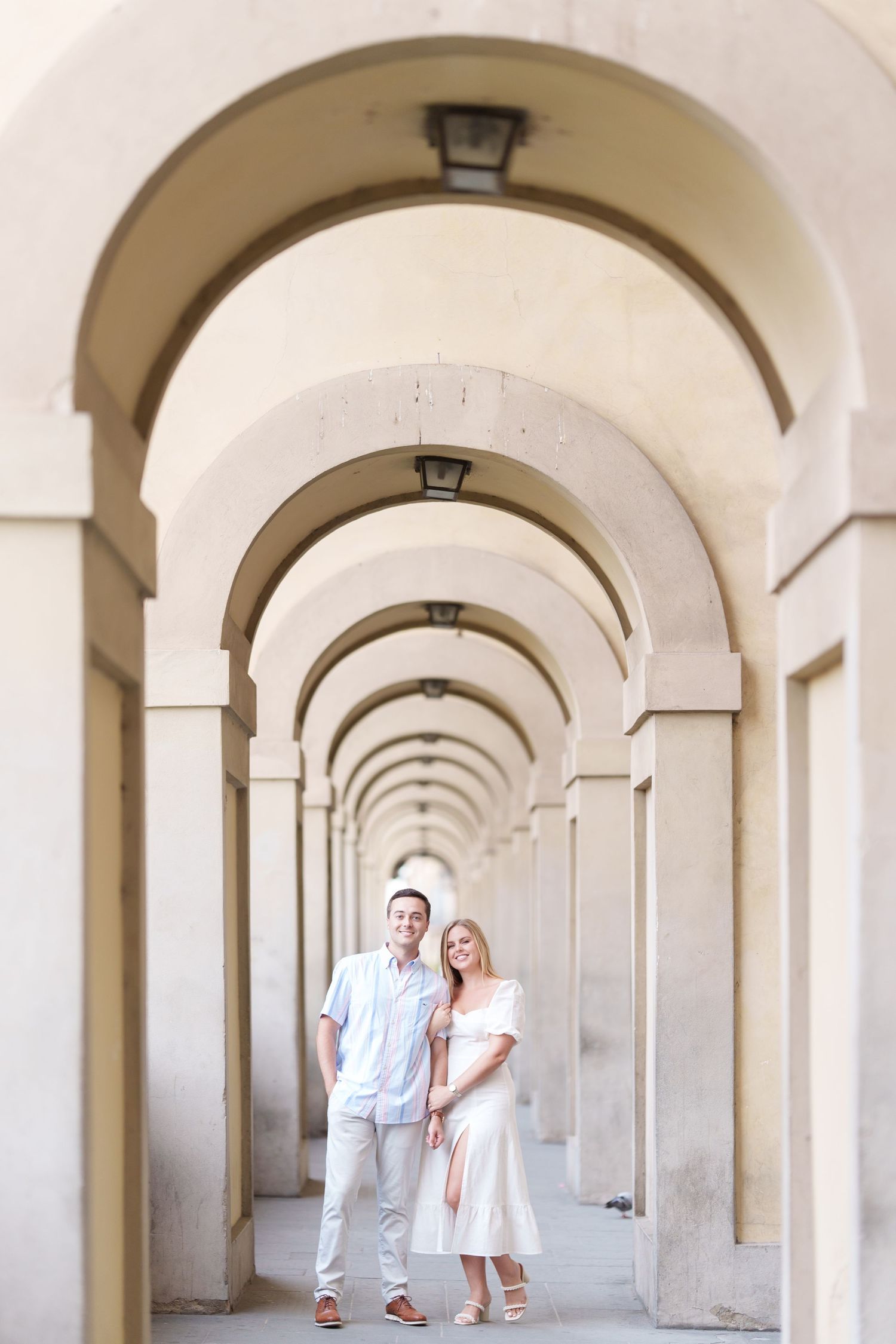 Couple in white outfits standing in a beautiful arched hallway with repeating architectural columns and natural light.