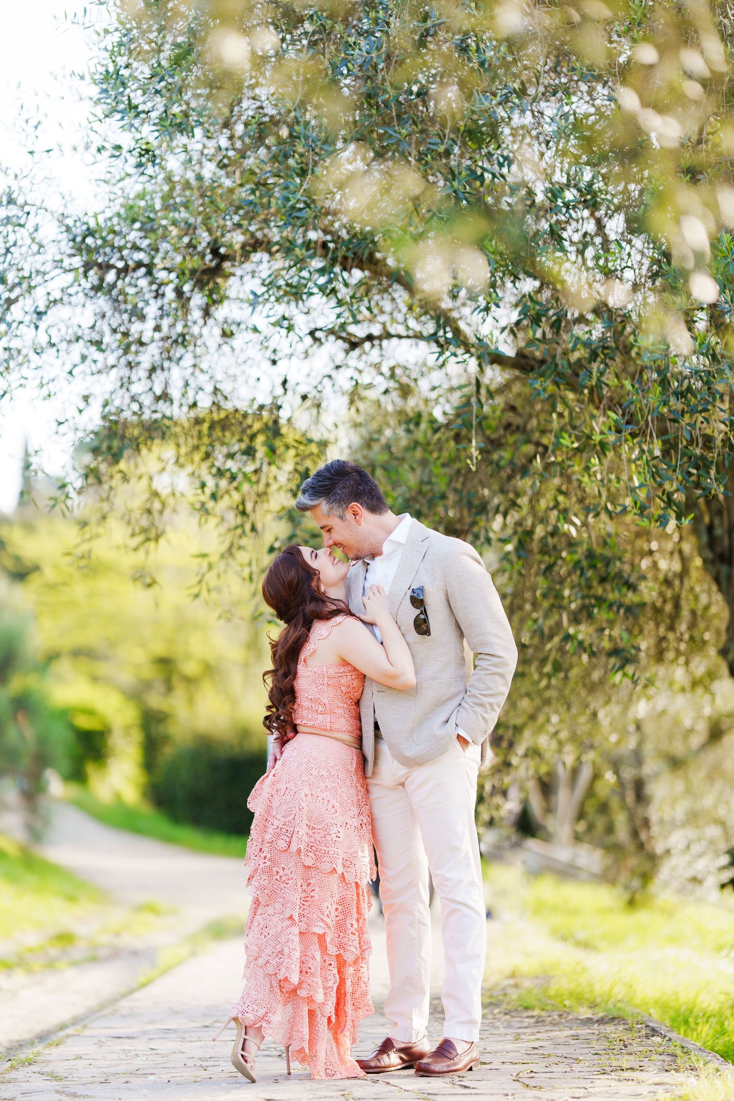 A couple in pink and white attire share a romantic moment under sunlit trees in a garden setting.