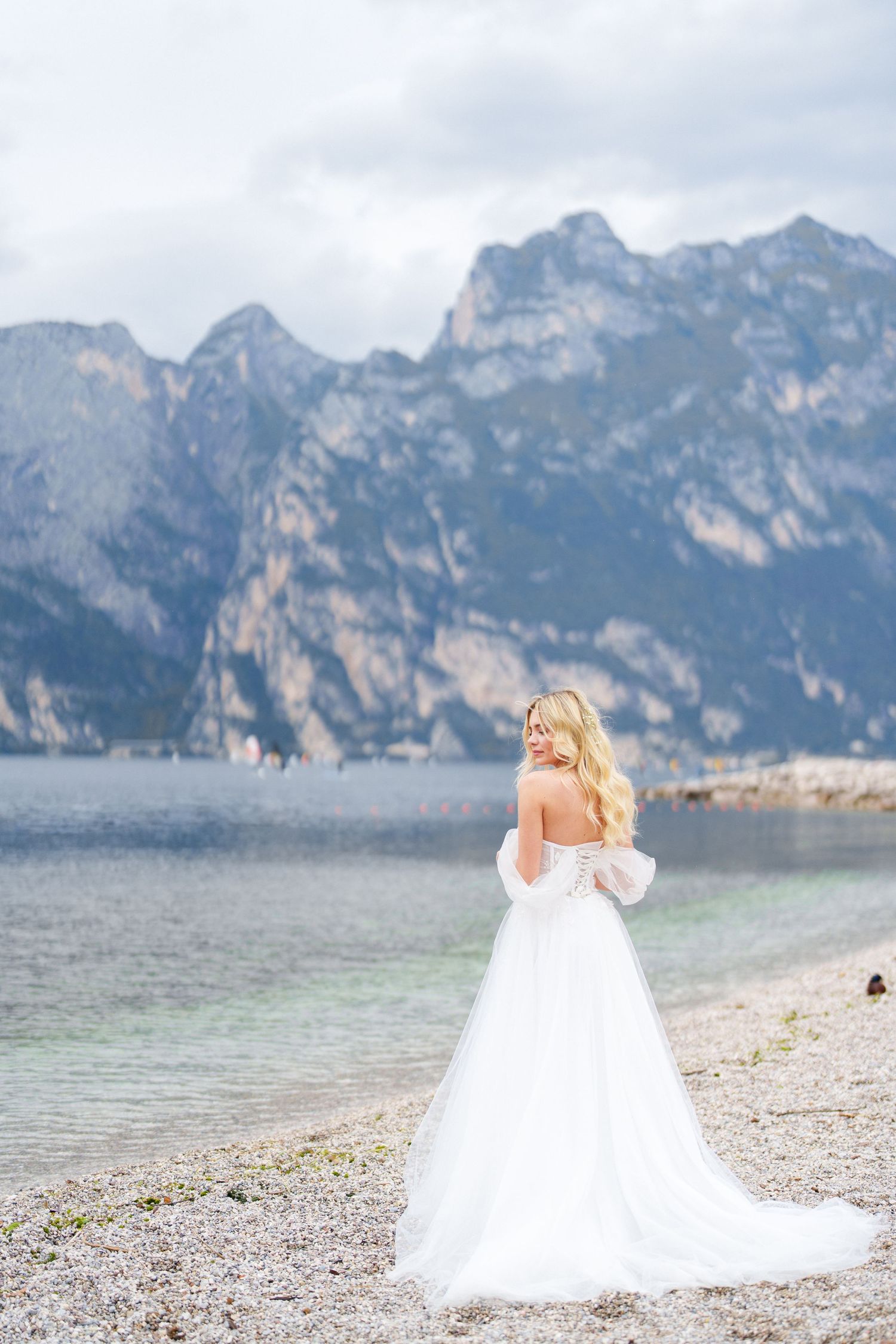 A bride in a flowing white gown stands on a rocky beach shore with dramatic mountain peaks rising over a serene lake.