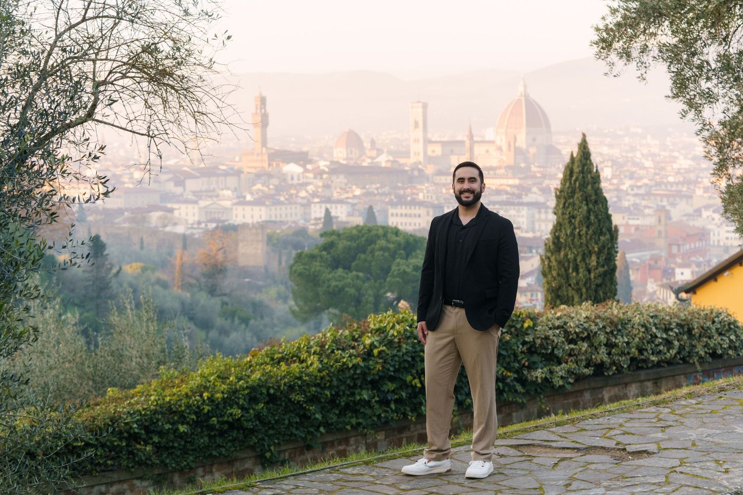 A scenic panoramic view of a city skyline from a hilltop path during sunset with trees and stone walkway.