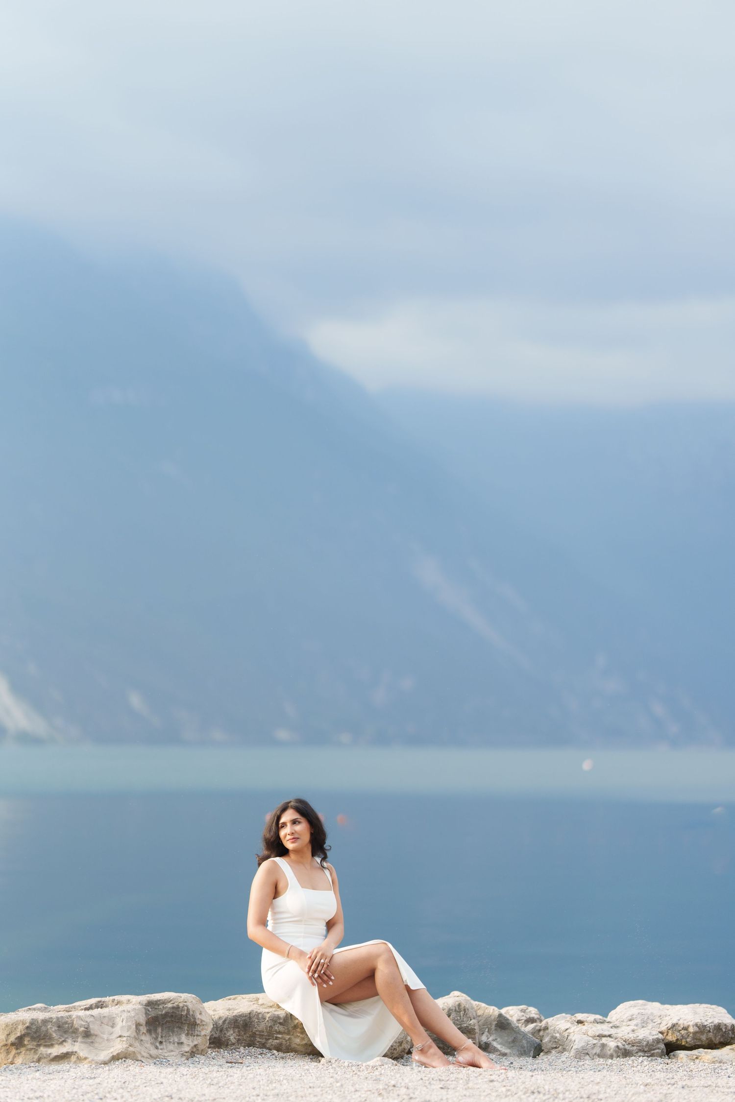 A person in white dress poses gracefully on rocky coastal cliffs overlooking a misty ocean and mountain landscape.