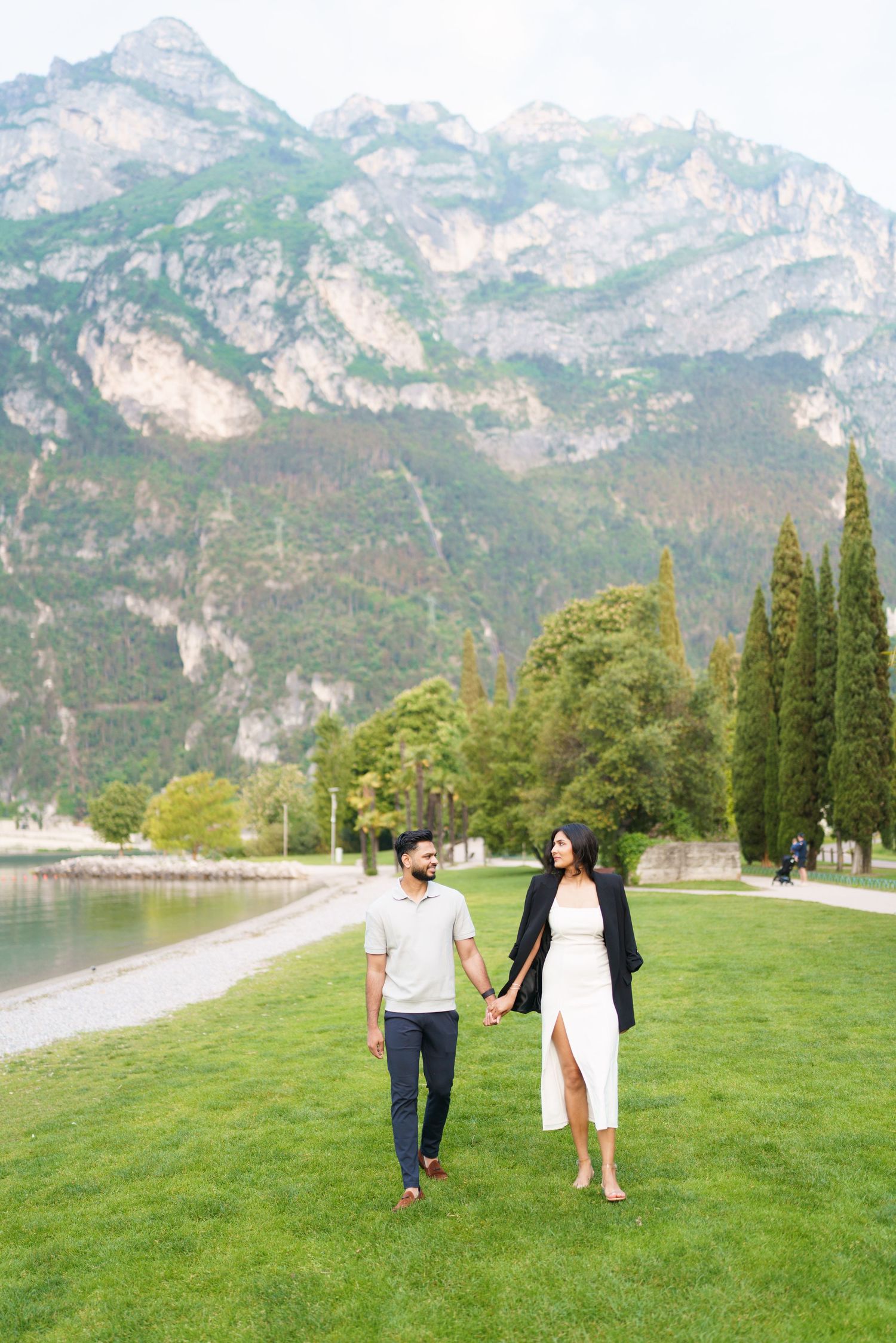 A couple walks hand in hand on lush green grass with majestic mountain peaks rising in the background near Lake Garda, Italy.