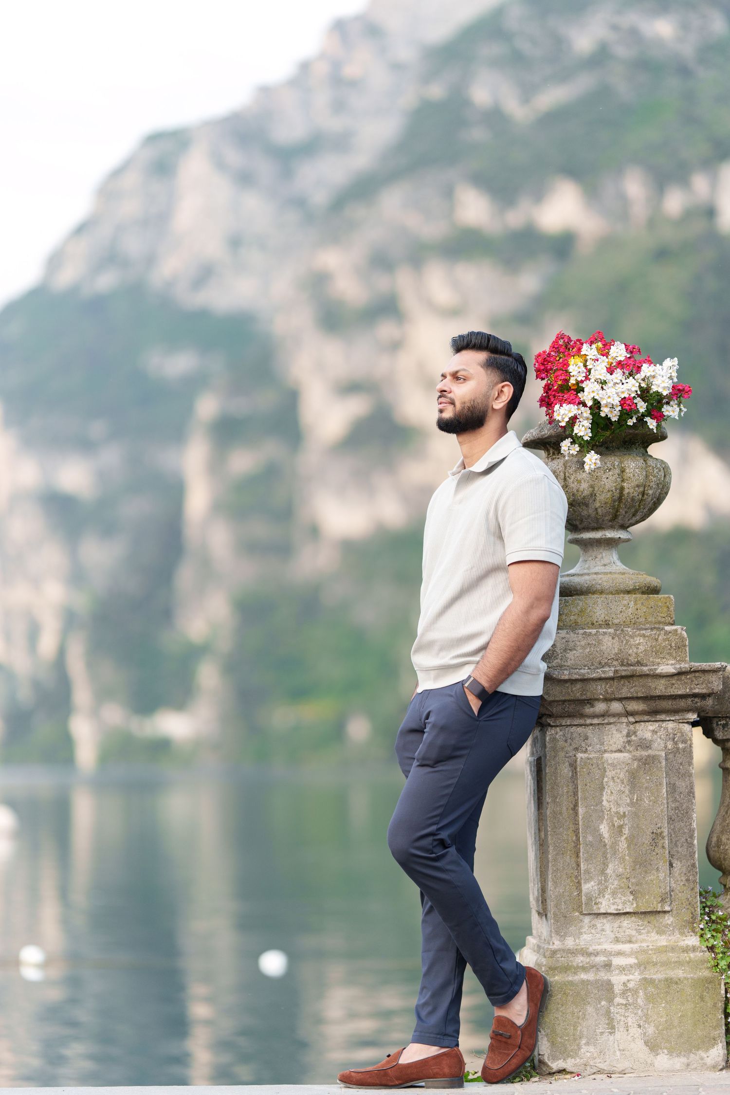 A person in casual attire poses by a stone pillar with flowers overlooking a scenic mountain lake landscape.