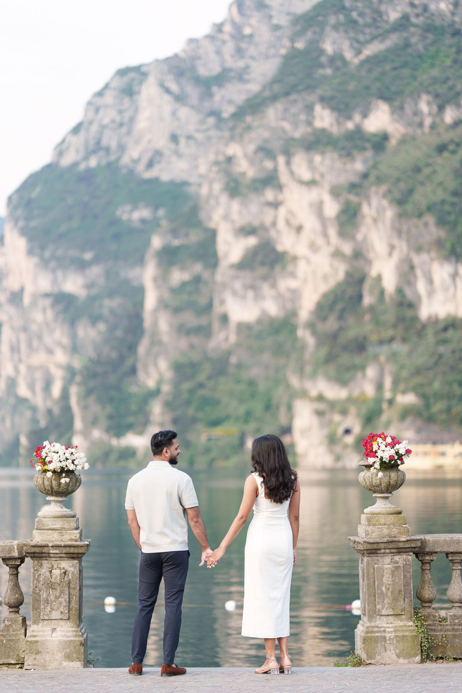 Couple holding hands on lakeside terrace with ornate stone pillars and dramatic mountain cliffs in background.