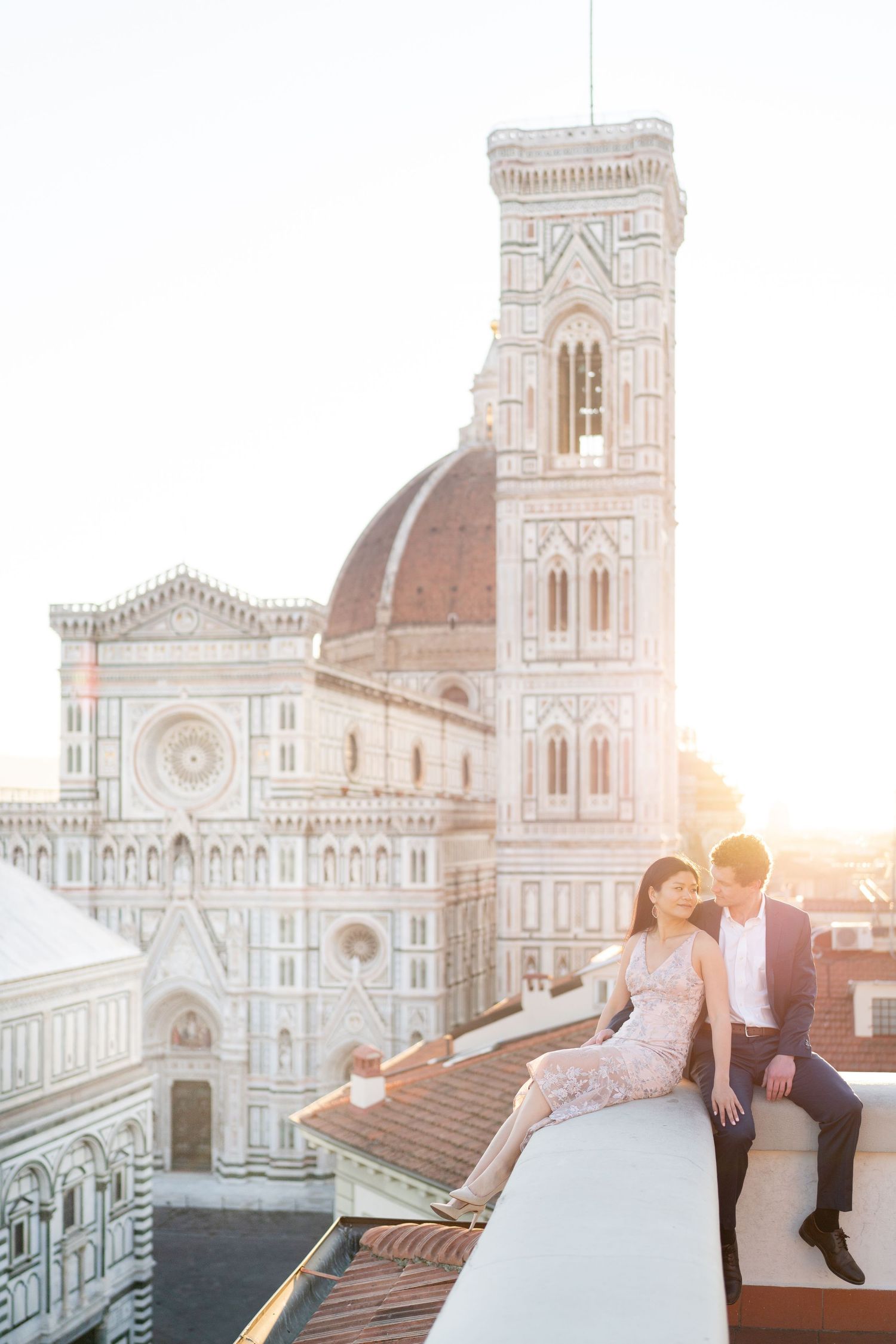 A couple sits on a rooftop overlooking the Florence Cathedral and Giotto's Bell Tower during golden hour sunset.