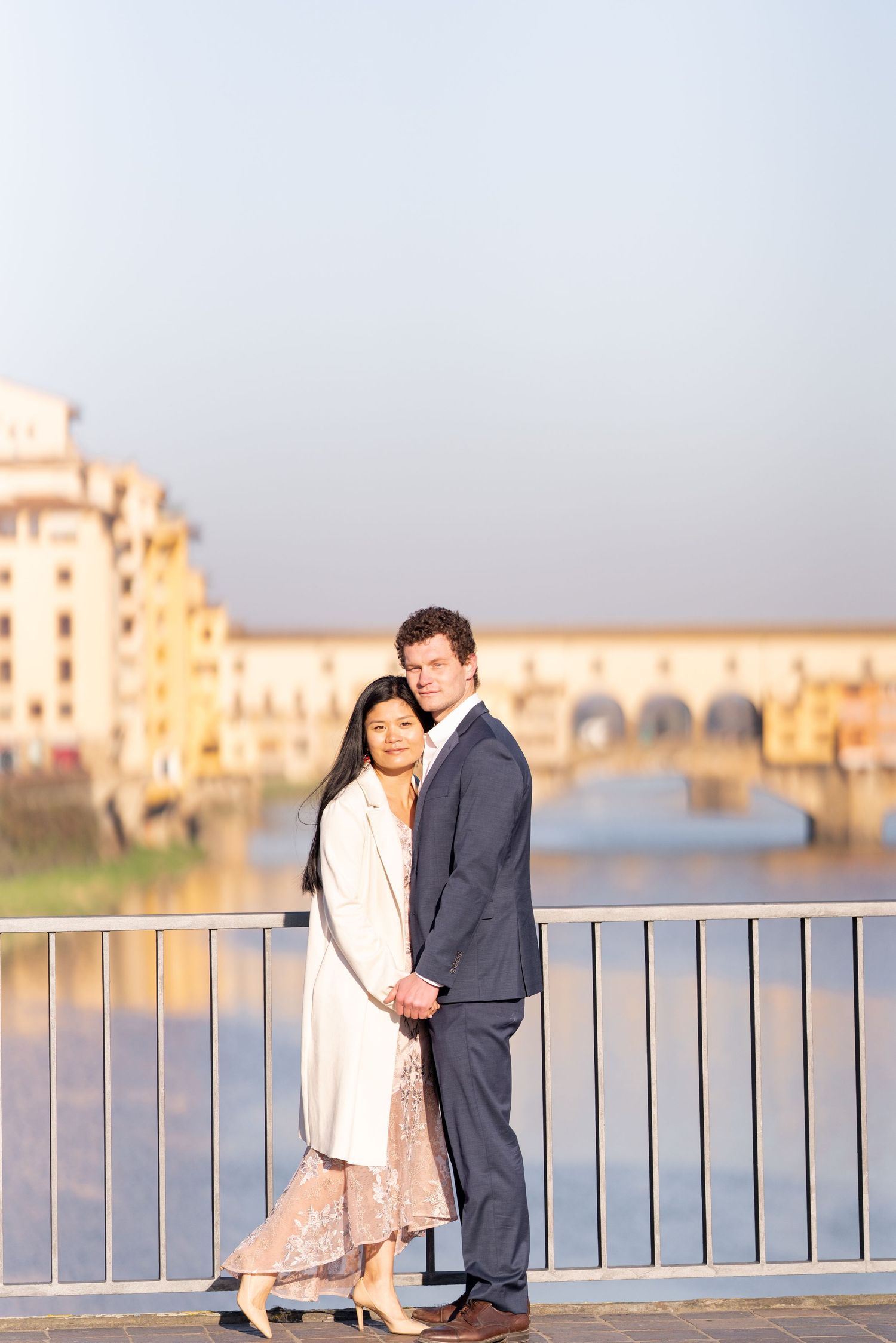 A romantic couple shares tender moments along a waterfront railing at sunset with urban buildings in the background.
