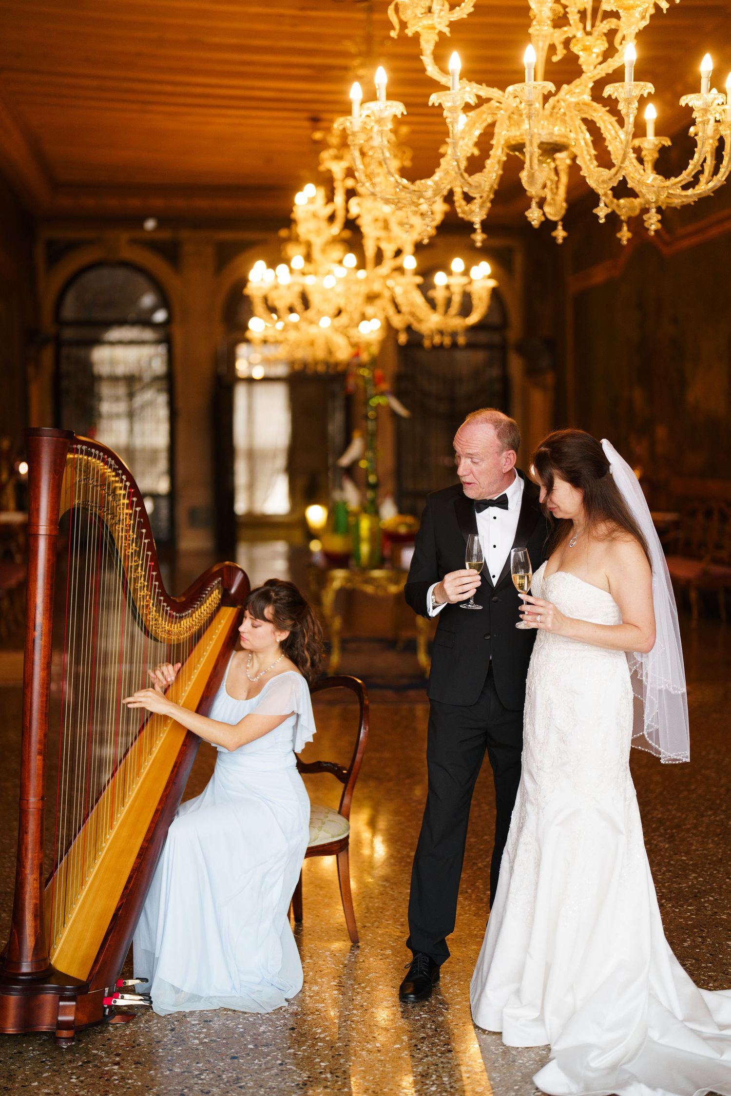 Elegant wedding couple dances together under ornate crystal chandeliers in a grand ballroom.