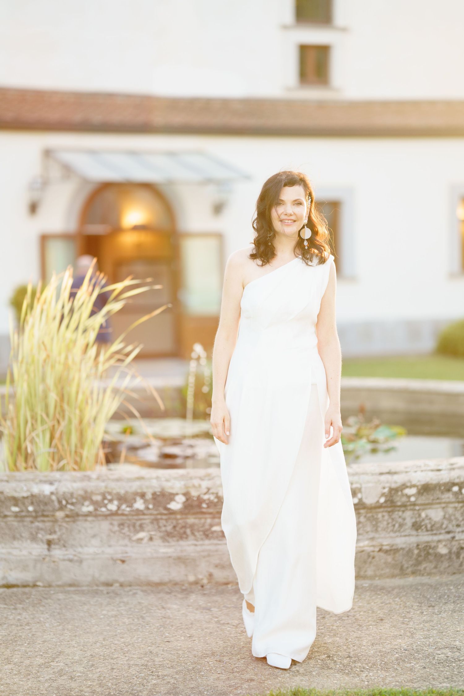 Flowing white wedding dress captured in sunlit outdoor setting near stone wall.