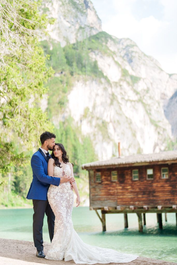 A wedding couple embraces near a wooden boathouse with majestic mountain peaks in the background.