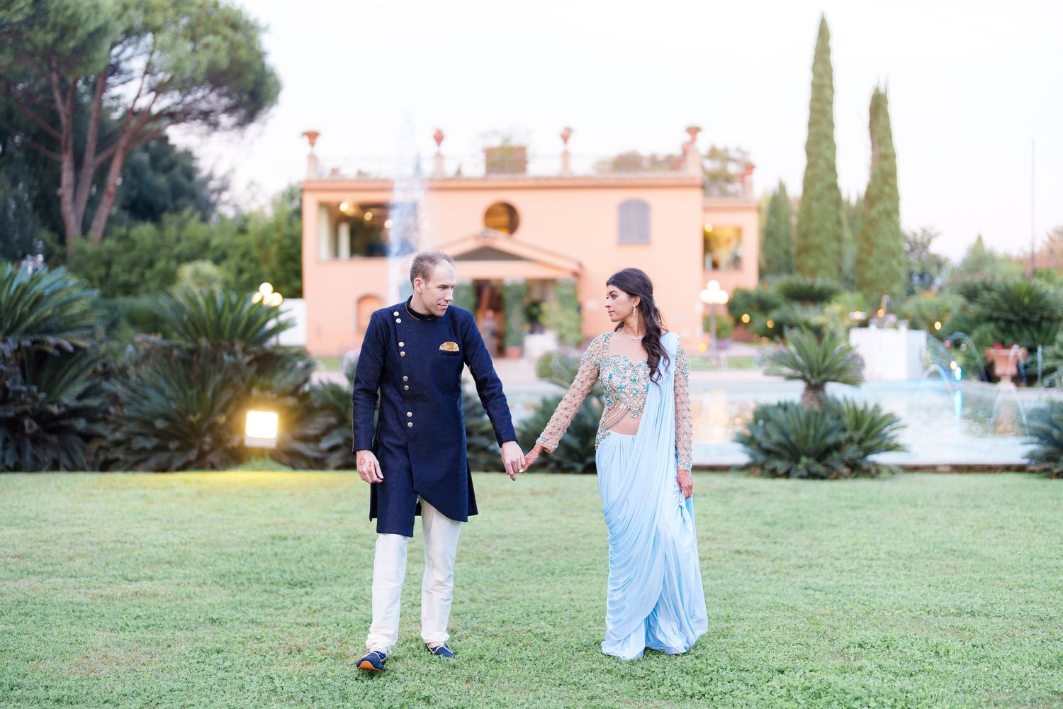 Couple in fusion wedding attire walking through garden with Mediterranean villa in background.