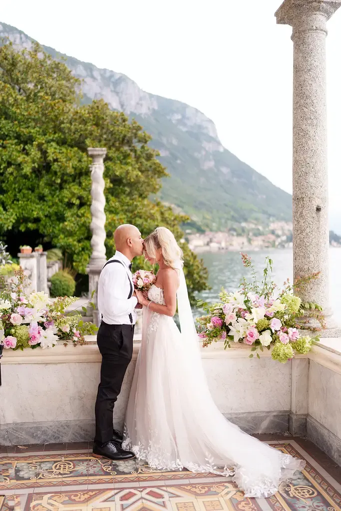 Wedding couple shares intimate moment on elegant terrace overlooking Italian lake and mountains.
