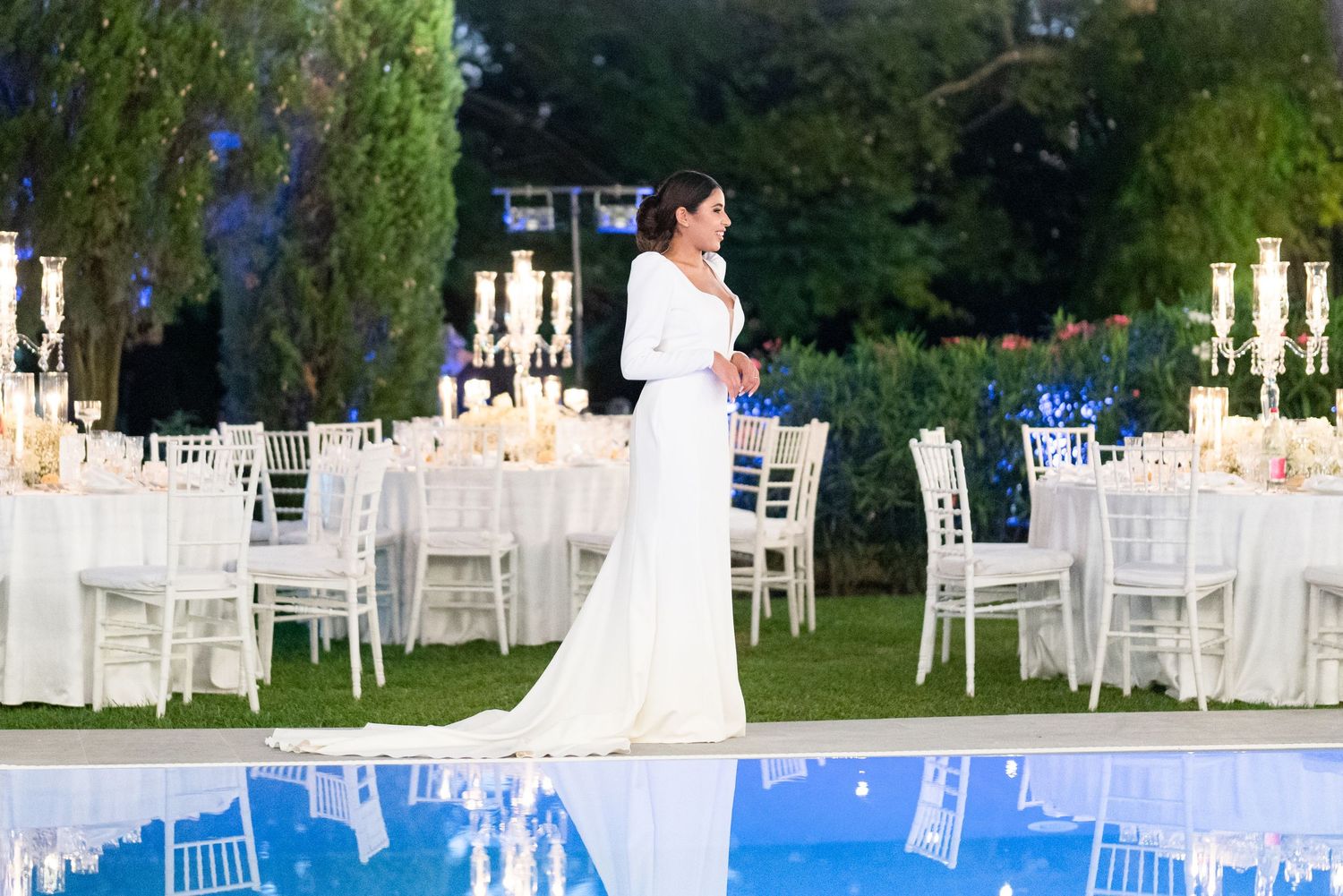 Outdoor wedding reception setup with white chairs and tables under string lights at dusk.