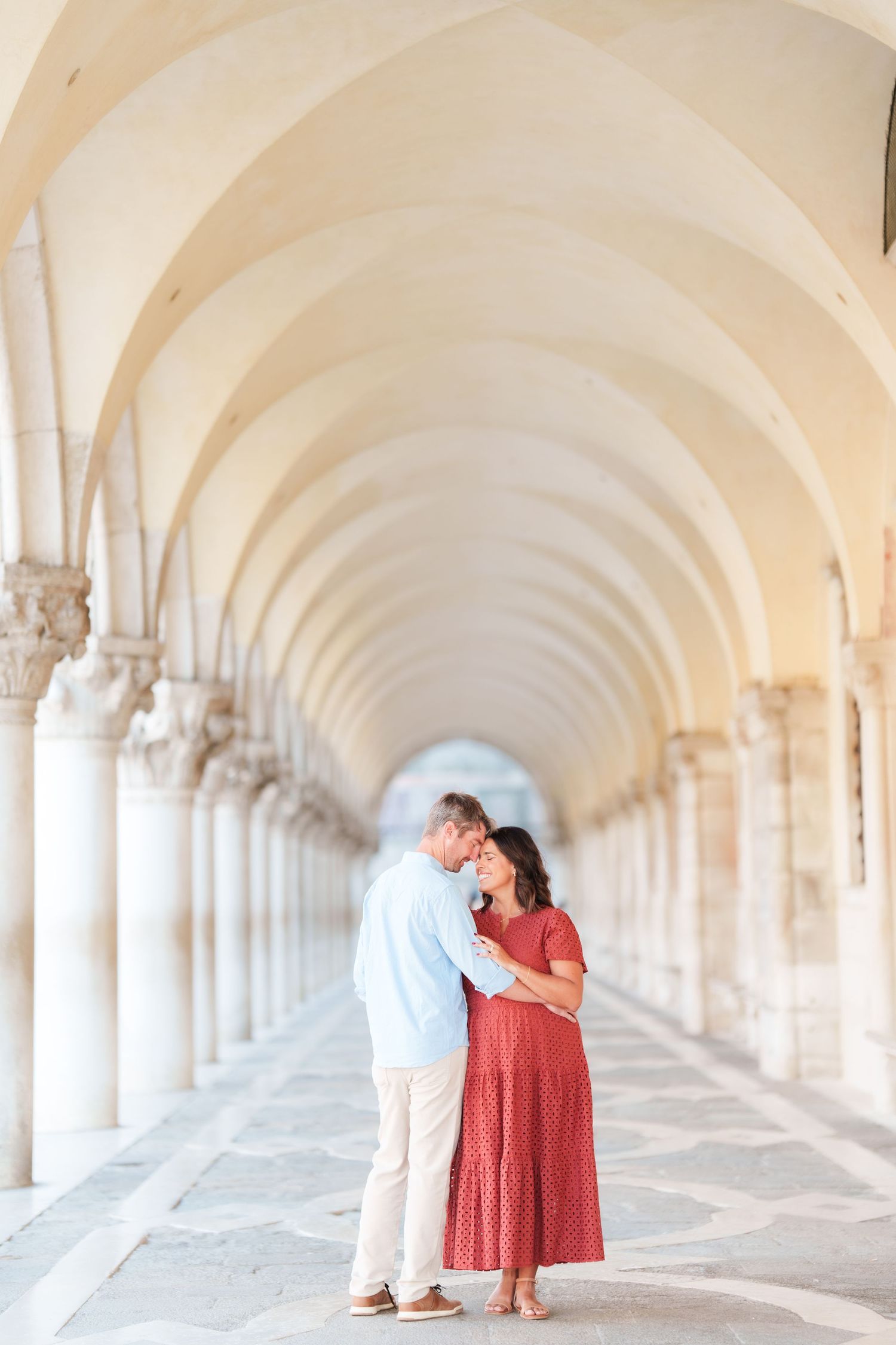 Couple embracing under elegant arched colonnade in Venice, woman in red dress, man in light blue shirt.