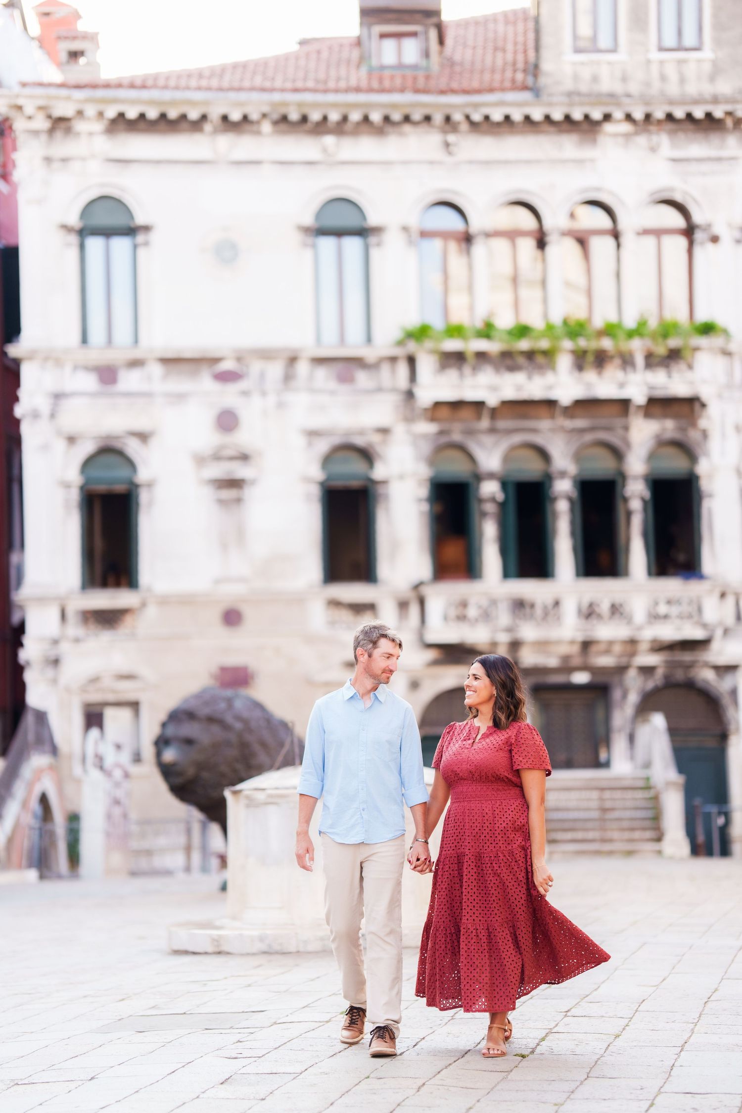 Couple holding hands and walking in a sunlit Venetian piazza with historic architecture in the background.