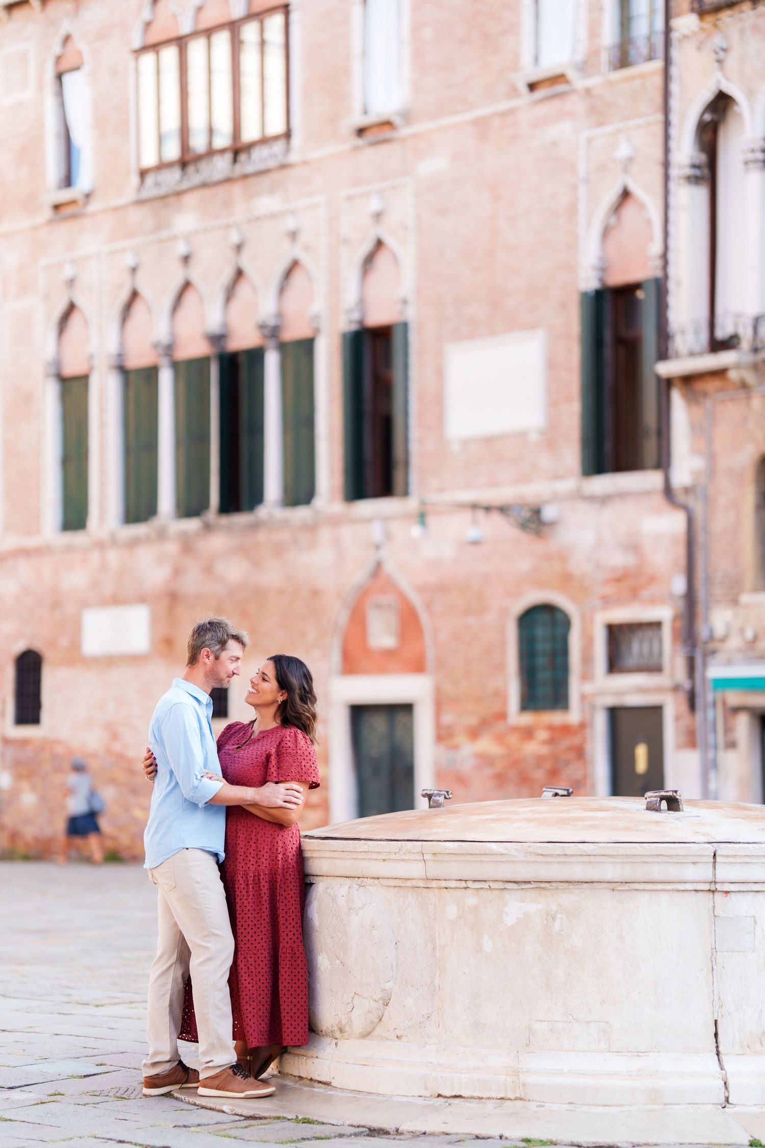 Couple embracing romantically by a stone fountain in a historic Venetian piazza with classic architecture.