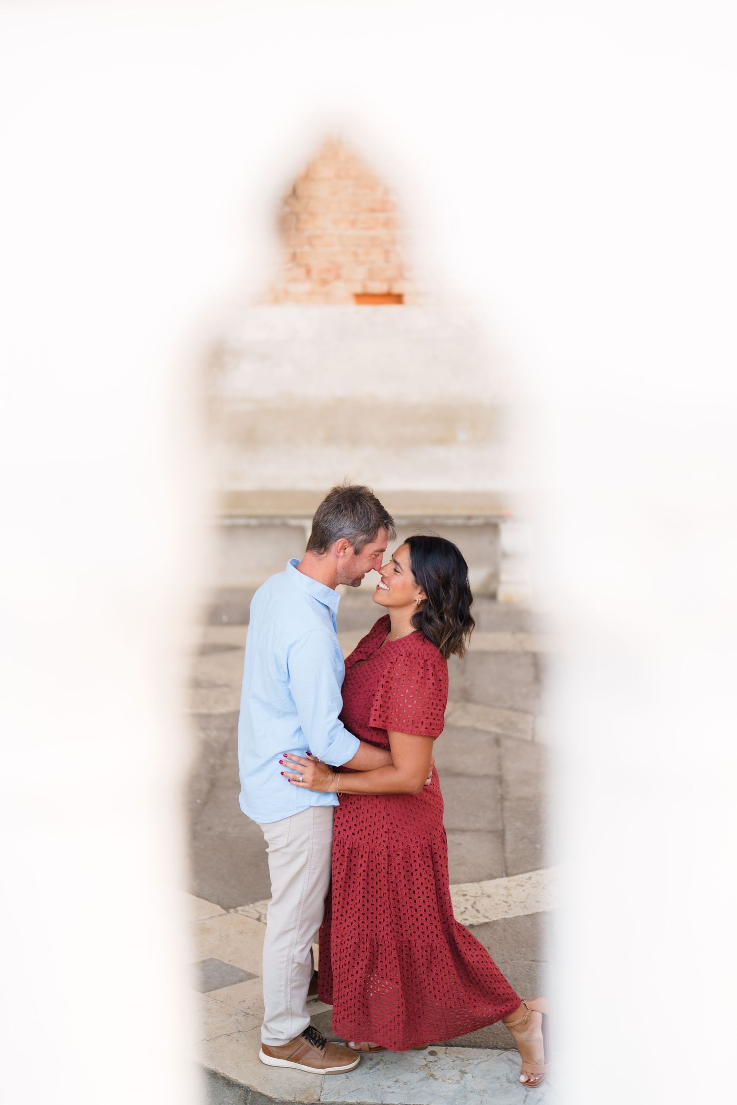 Romantic couple embracing and almost kissing, framed by a white architectural arch, woman in red dress, man in light blue shirt.
