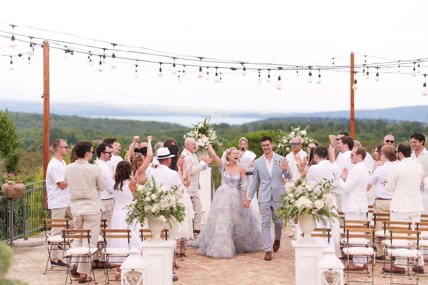 Outdoor wedding ceremony with bride in blue gown, guests in white, string lights, and scenic hilltop views.