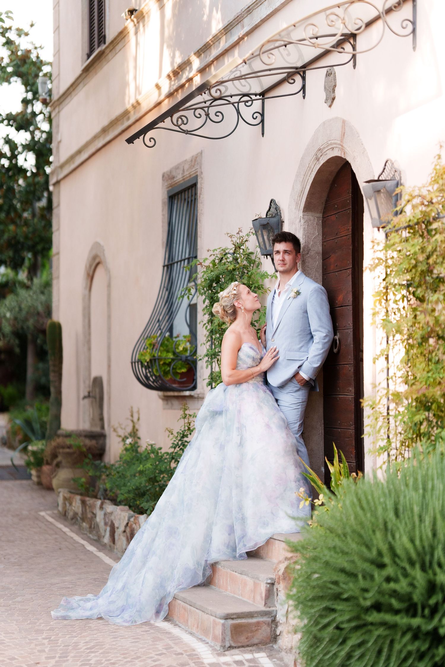 Bride in blue floral gown and groom in gray suit pose on steps outside elegant Mediterranean villa.
