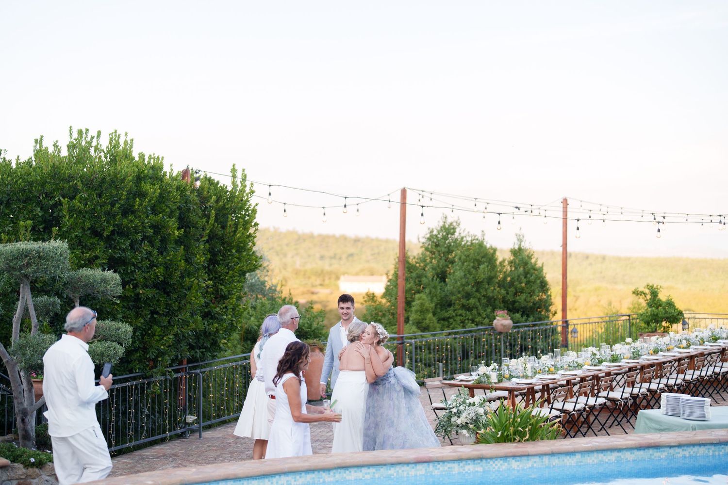 Outdoor wedding ceremony by a pool with string lights, lush greenery, and a scenic countryside backdrop at sunset.