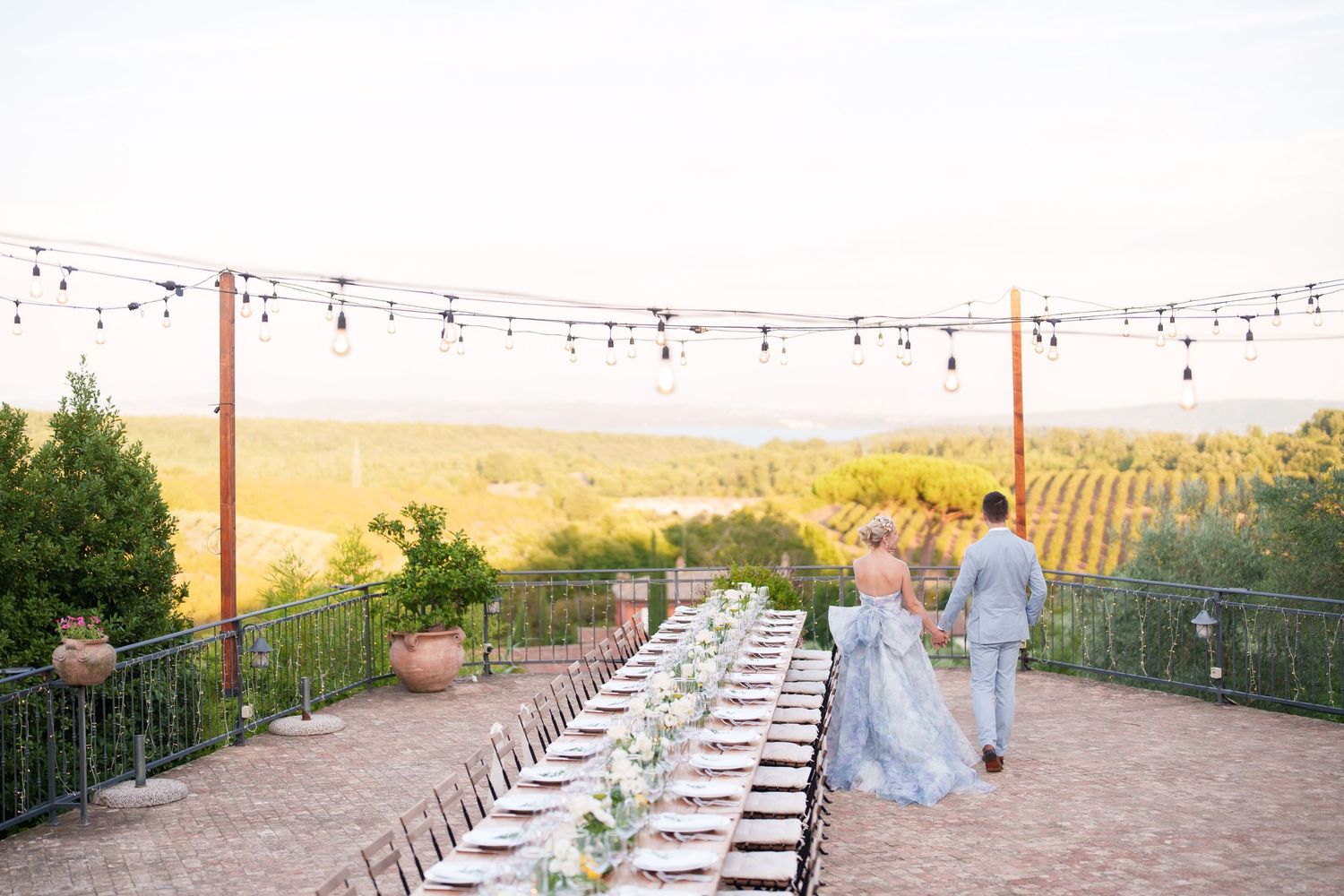 Bride and groom walk past long outdoor reception table with string lights overlooking vineyard at sunset.