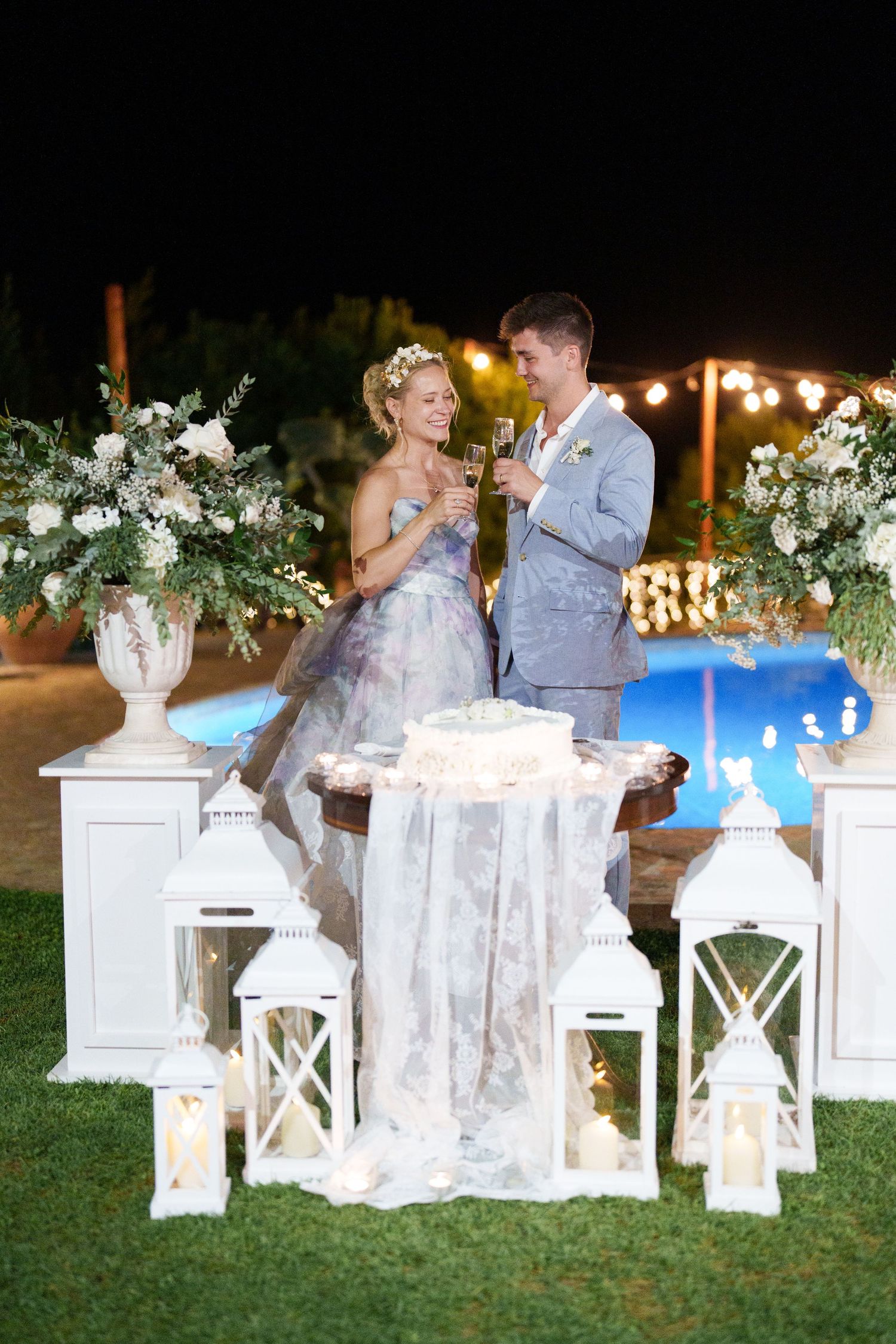 Bride and groom toasting champagne at outdoor night wedding reception with white lanterns and floral arrangements.