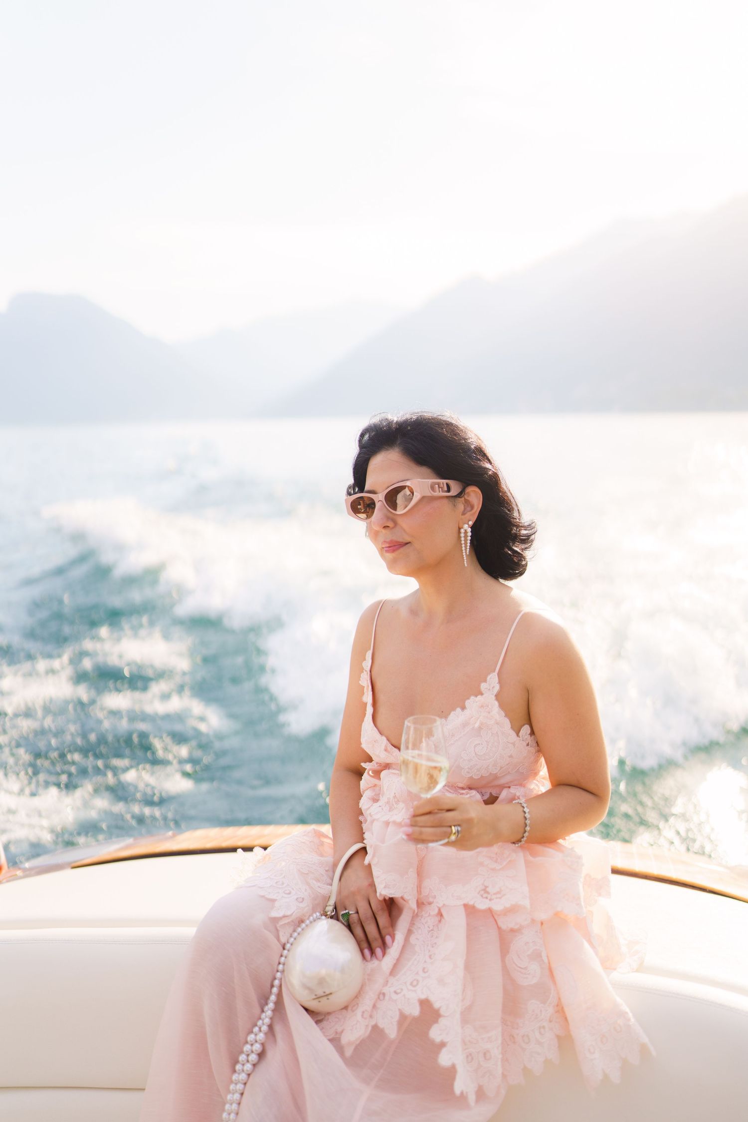 Elegant woman in pink lace dress holding champagne on a luxury yacht with mountain backdrop.