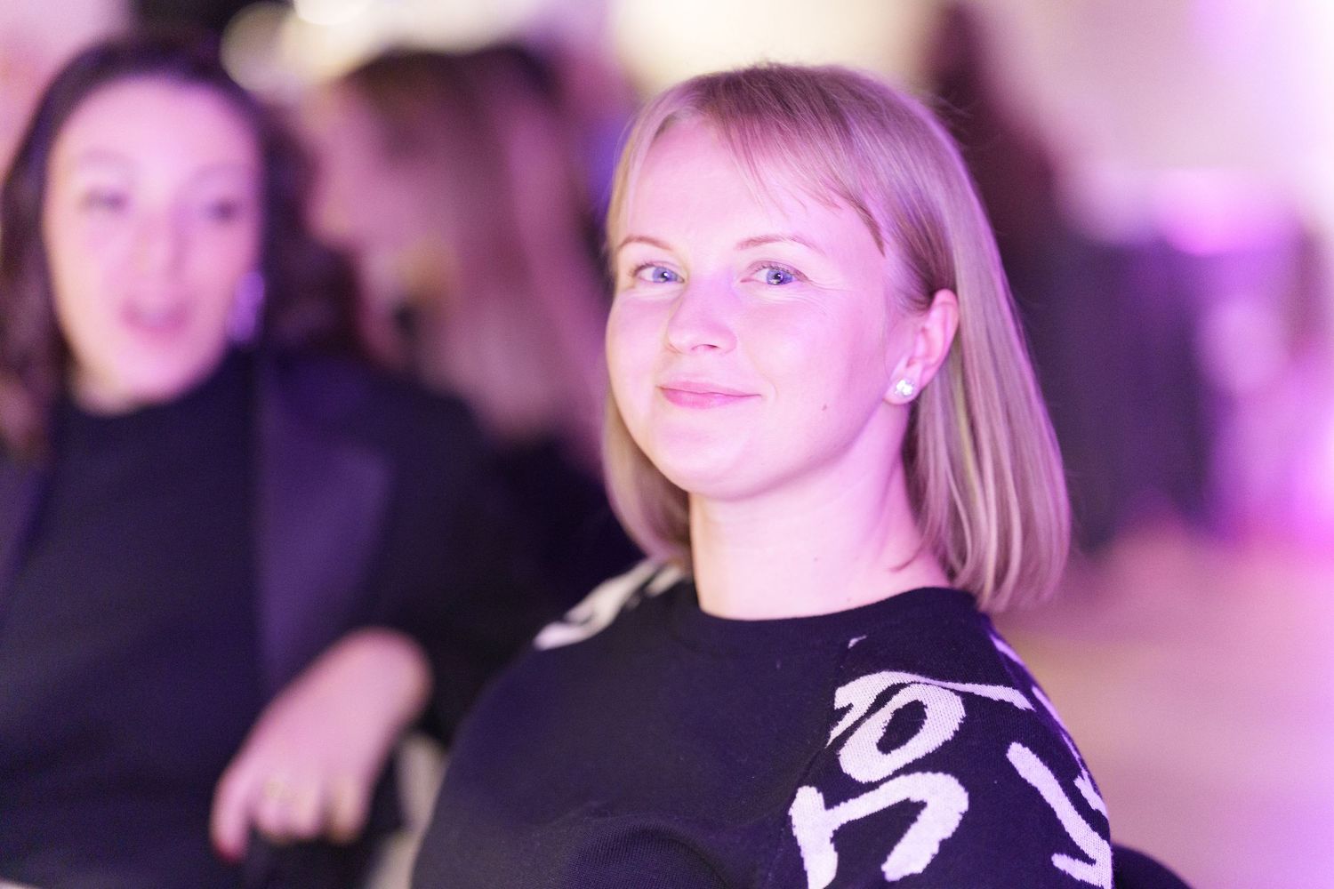Young blonde woman in black top smiling at an event with purple lighting in the background.