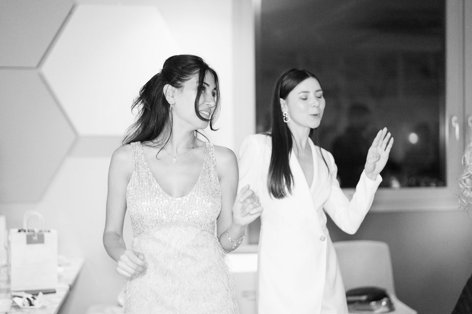 Black and white photo of two women in elegant dresses, one in a lace gown, sharing a moment indoors.